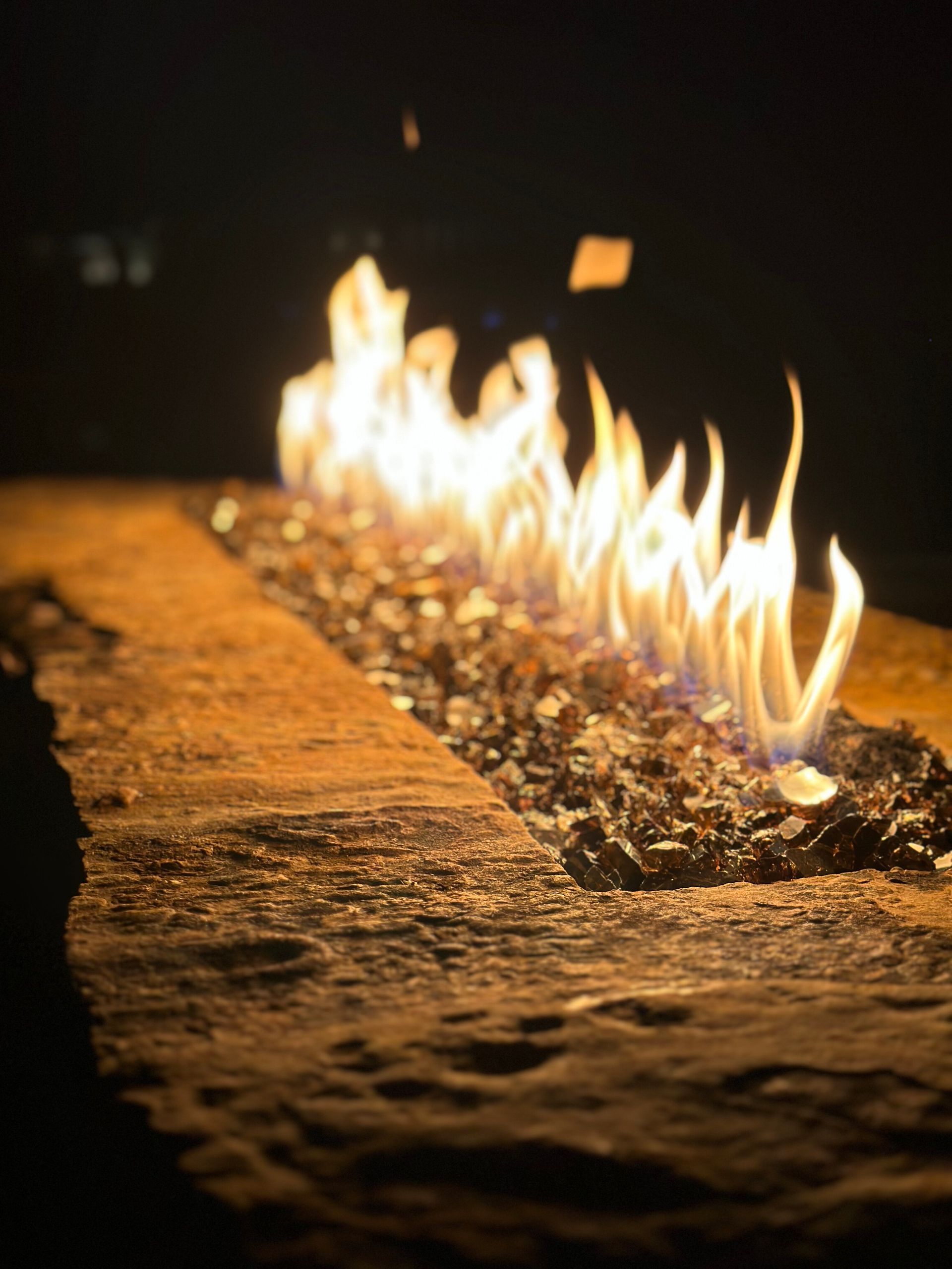 A linear outdoor fire pit with a bright, flickering flame emerging from a bed of dark glass rocks at night.