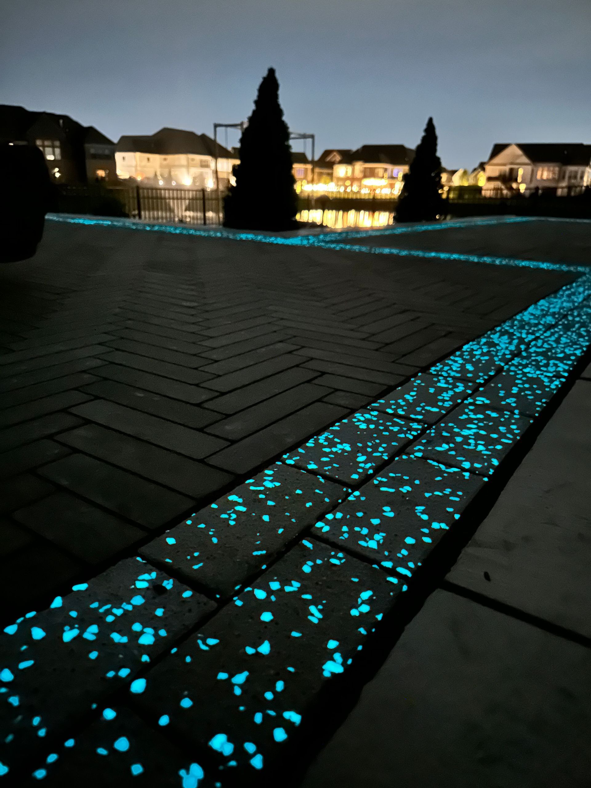 A paved walkway at night with glowing blue stones embedded in the border, illuminated against a blurred house background.
