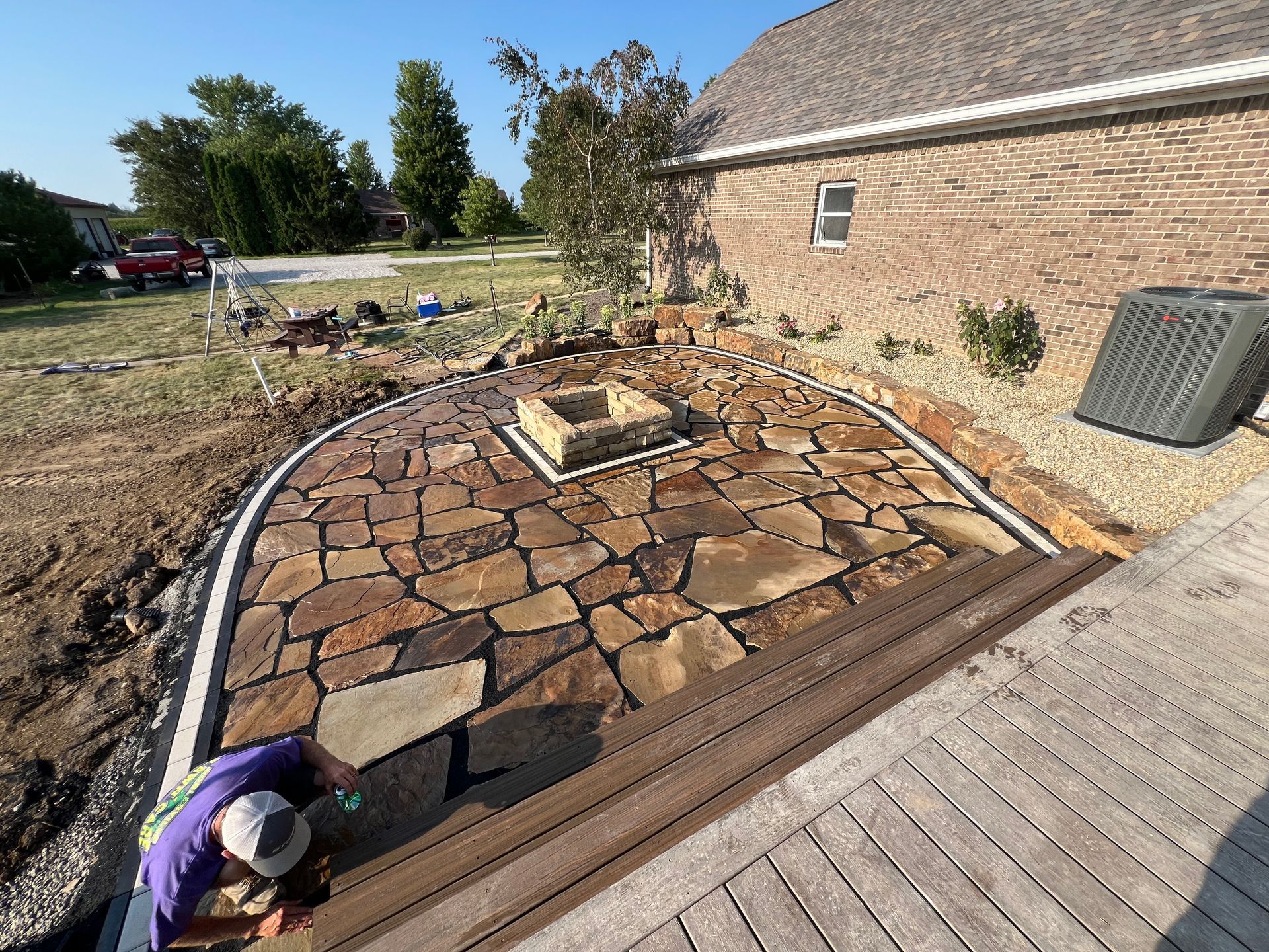 A worker installing a stone patio with a central fire pit next to a house and a wooden deck.
