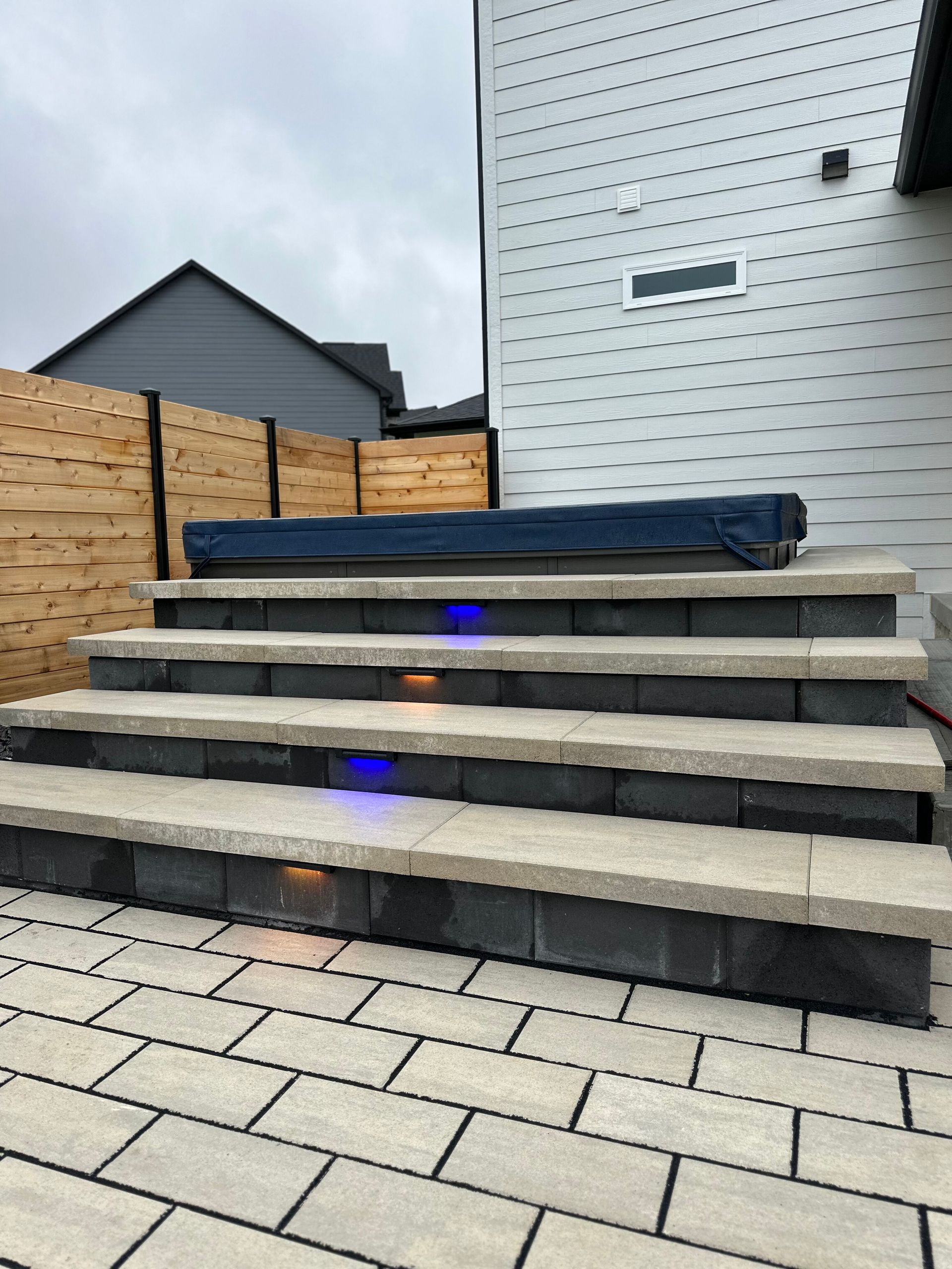 A hot tub with a dark blue cover sits atop wide, tiered stone stairs with integrated blue and warm accent lighting.