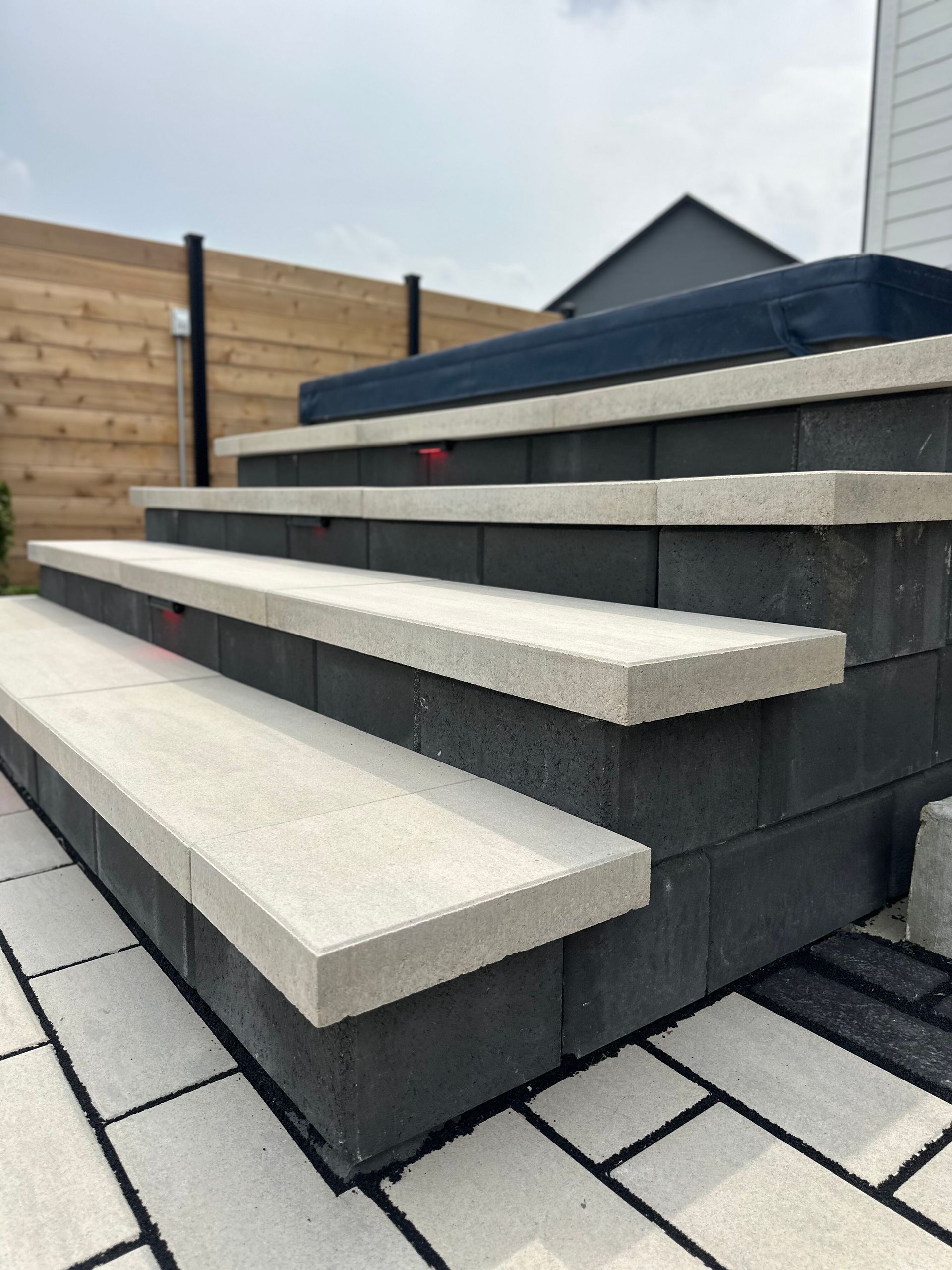 Outdoor steps with dark grey block risers and light grey stone treads leading up to a hot tub on a paved patio.