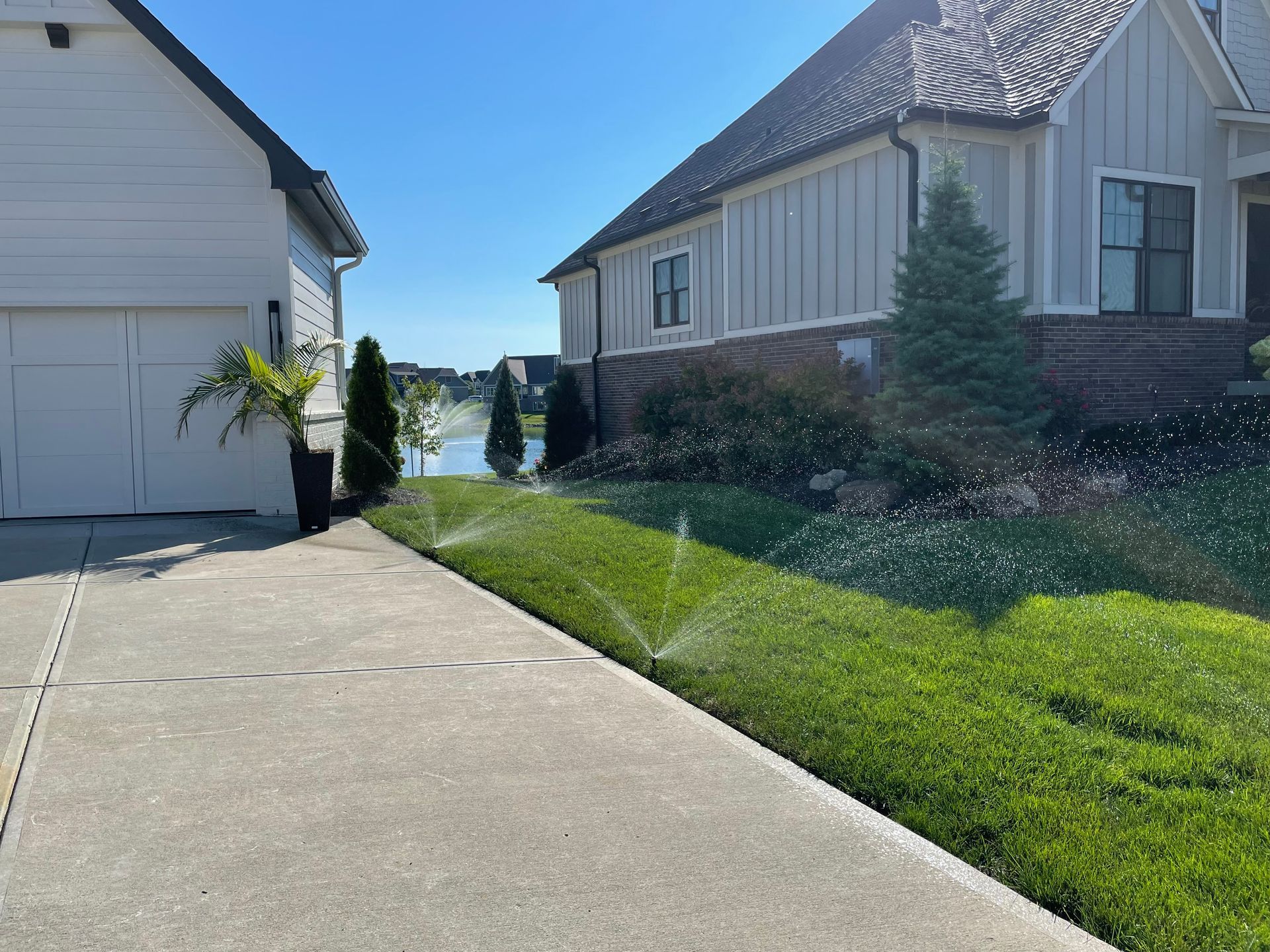 Sunlit residential lawn with sprinklers watering the grass between a white garage and a modern house.