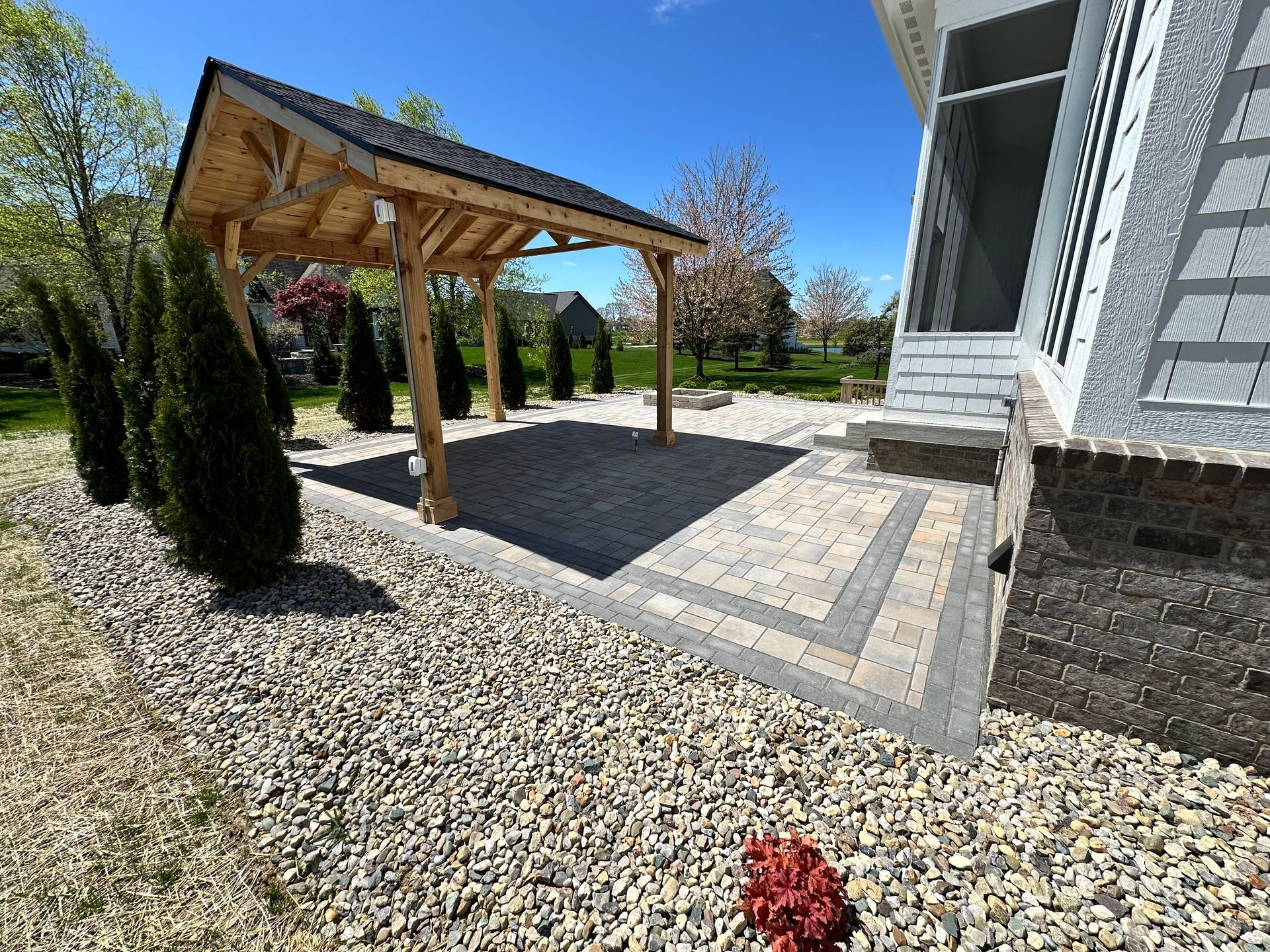 A wooden pavilion on a paver patio beside a house, surrounded by gravel landscaping and small evergreen trees.