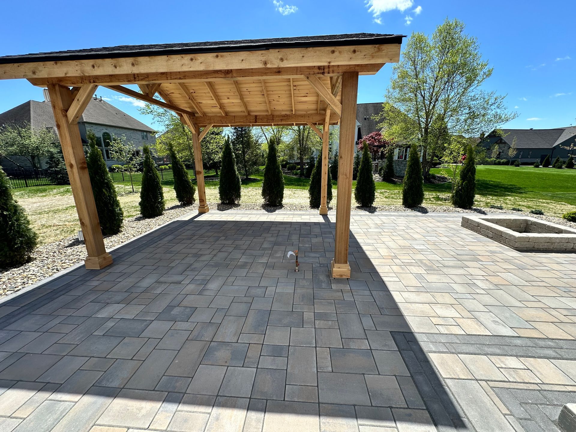 A wooden pergola over a gray stone patio in a sunny backyard, featuring a paved fire pit area and evergreen trees.