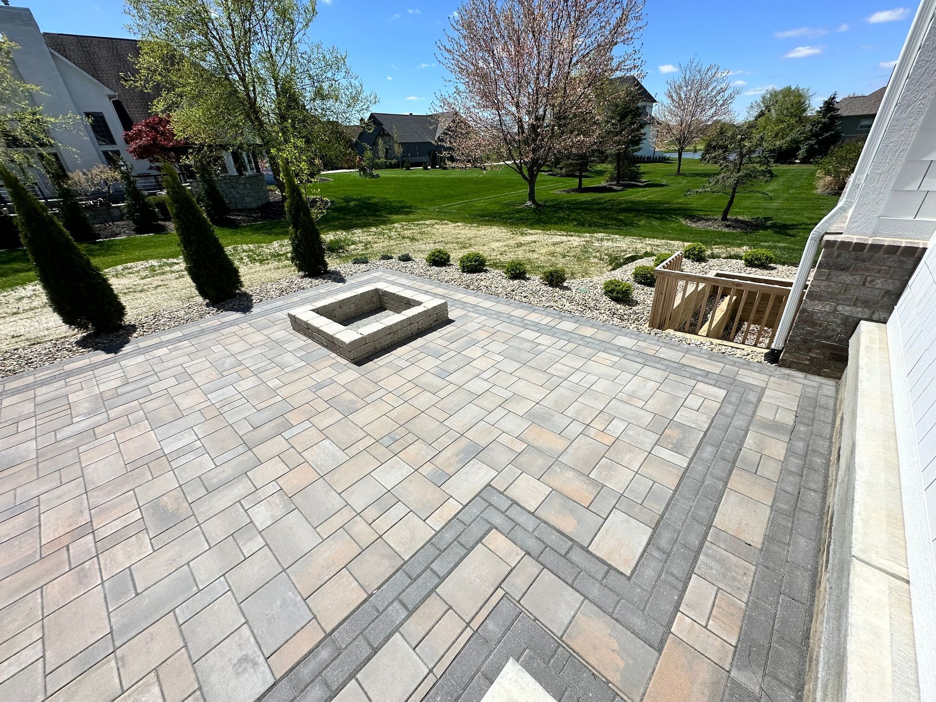 A backyard stone paver patio featuring a square fire pit, surrounded by landscaped lawn and trees on a sunny day.