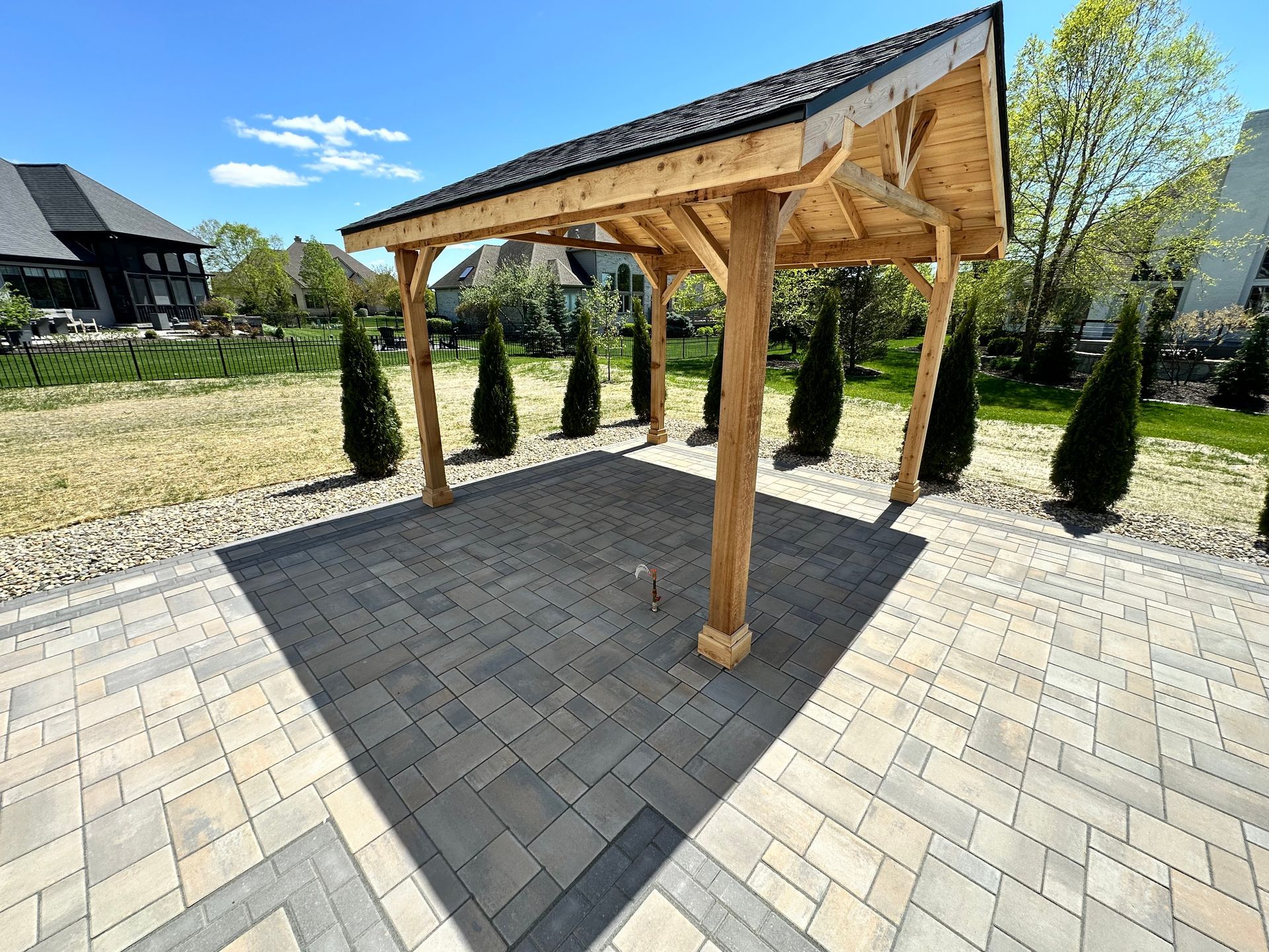 A wooden pergola stands on a paved stone patio in a sunny backyard, surrounded by small evergreen trees.