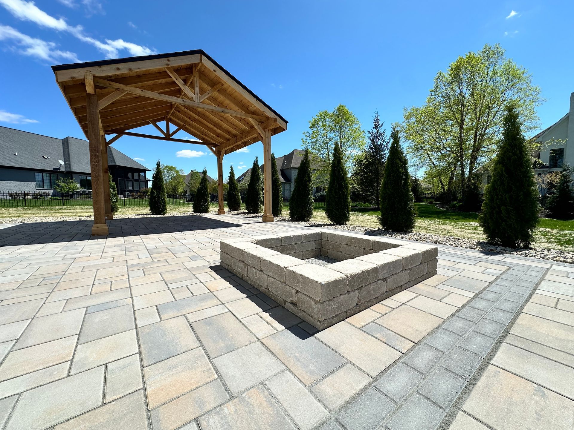 A stone patio featuring a square fire pit and a wooden pavilion under a clear blue sky, surrounded by evergreen trees.