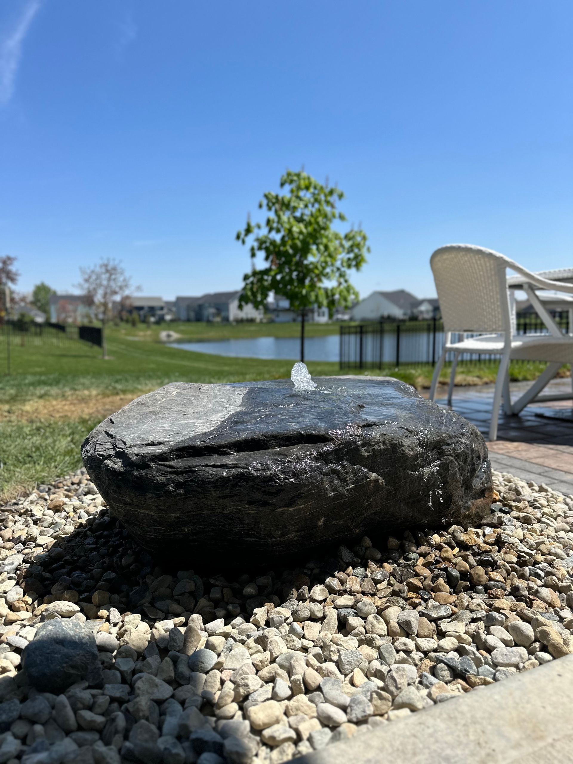 A stone water fountain feature sits in a gravel bed with a pond, tree, and patio chair in the background.