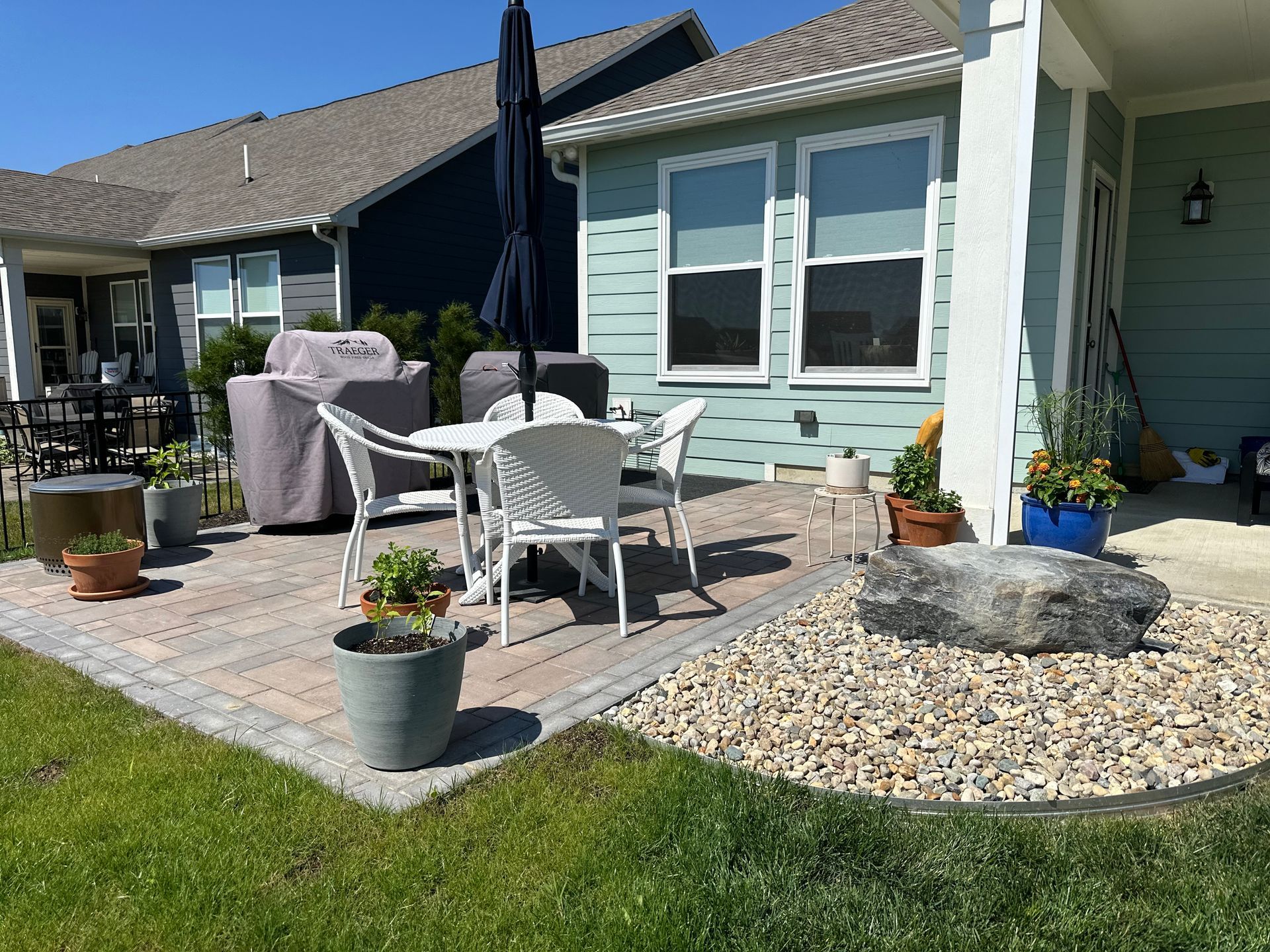 A light blue house patio with a dining set, gray grill, potted plants, and a landscaping rock on a gravel bed.