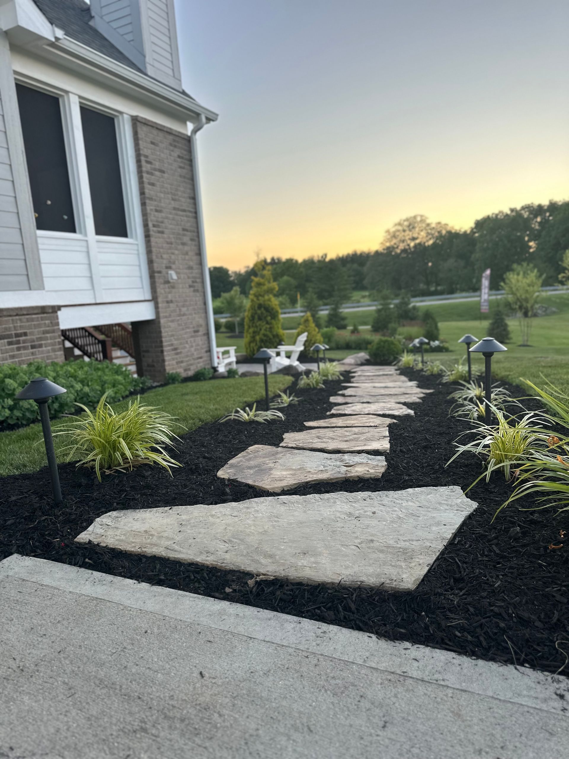 Stone stepping path through black mulch leading from a house patio toward a grassy lawn at sunset.