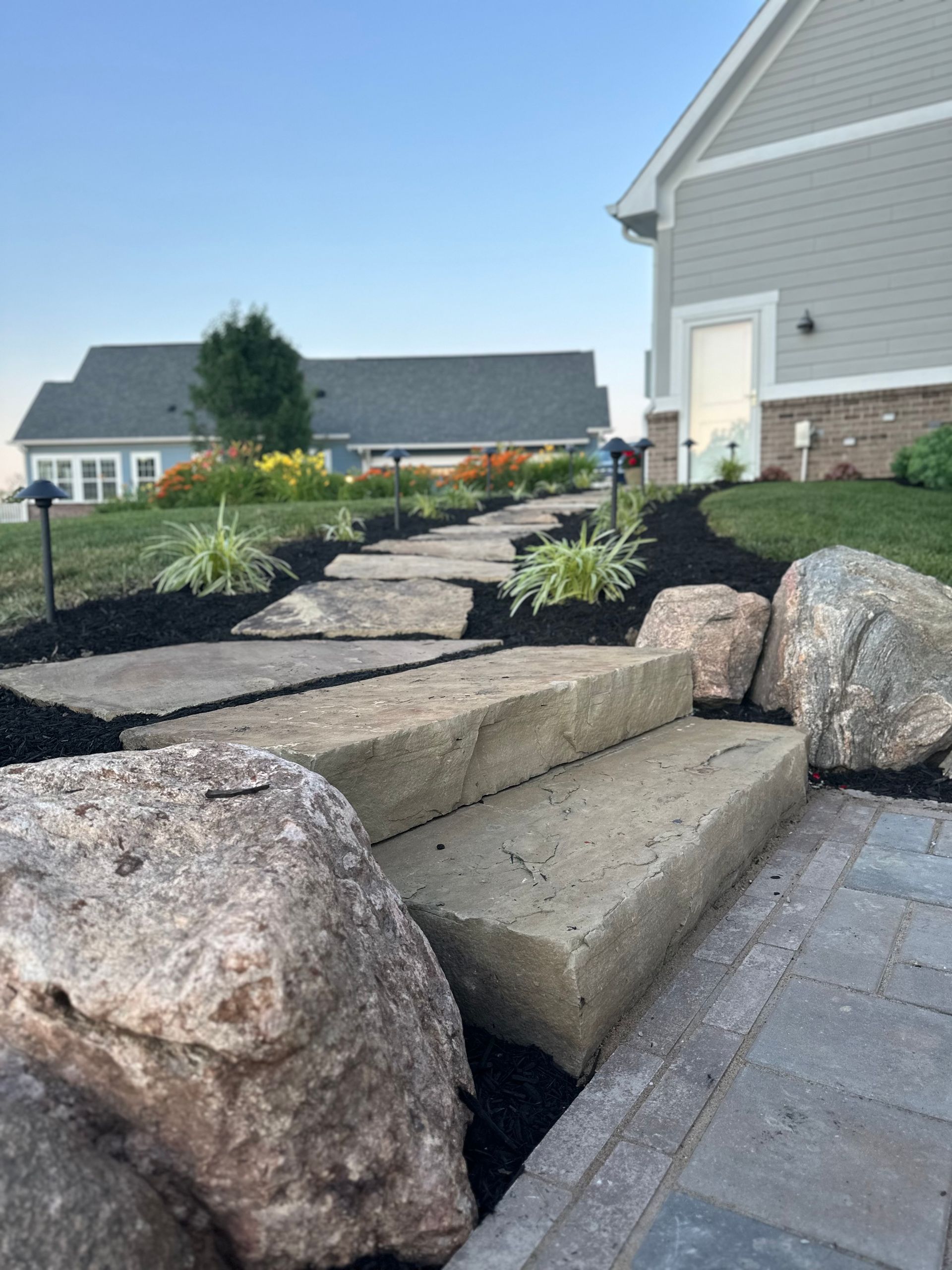 Concrete steps leading up a mulched landscaping path flanked by large boulders and small plants next to a house.