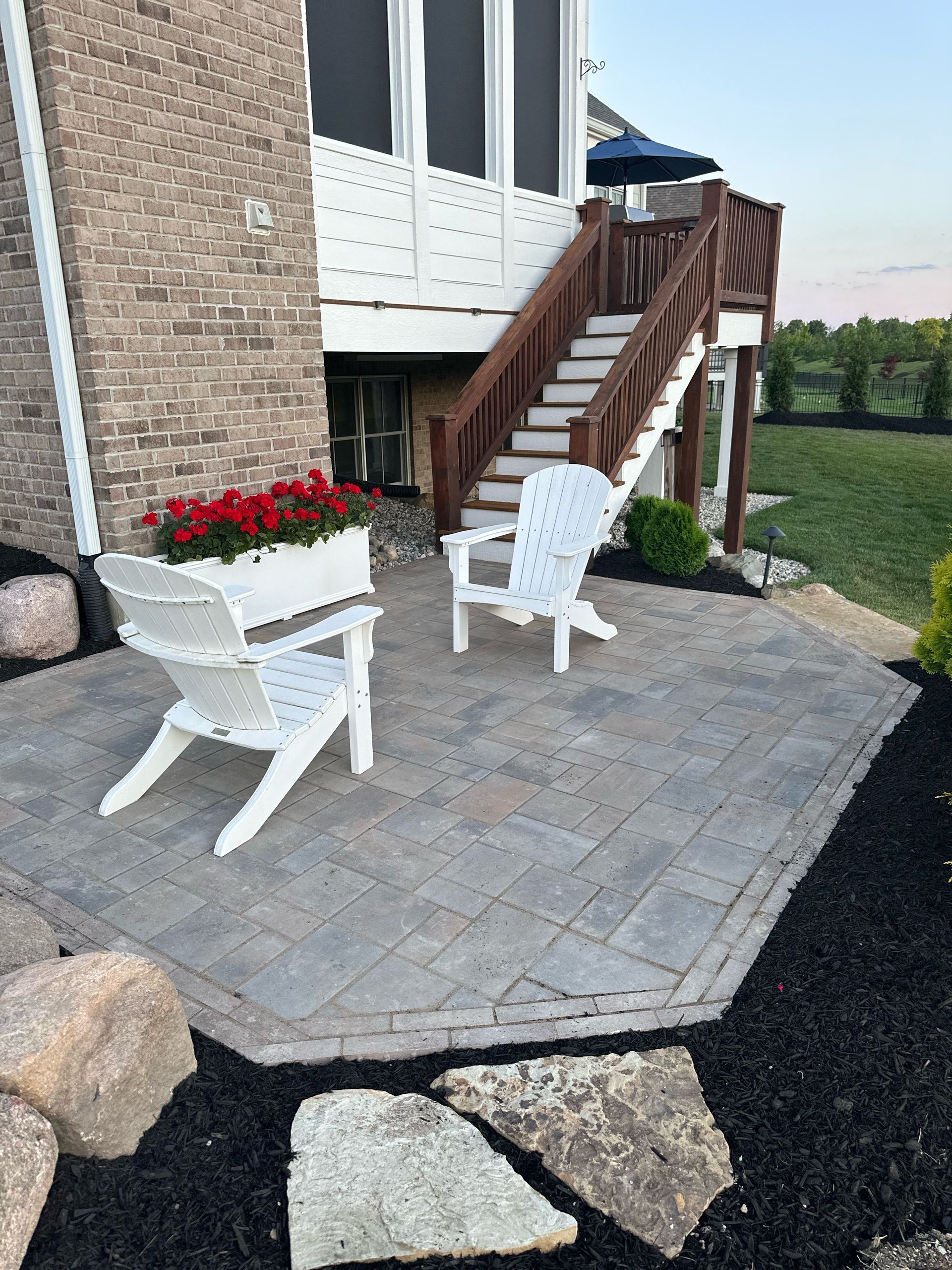 Two white Adirondack chairs on a grey paver patio next to a brick house with a wooden staircase and red flowers.