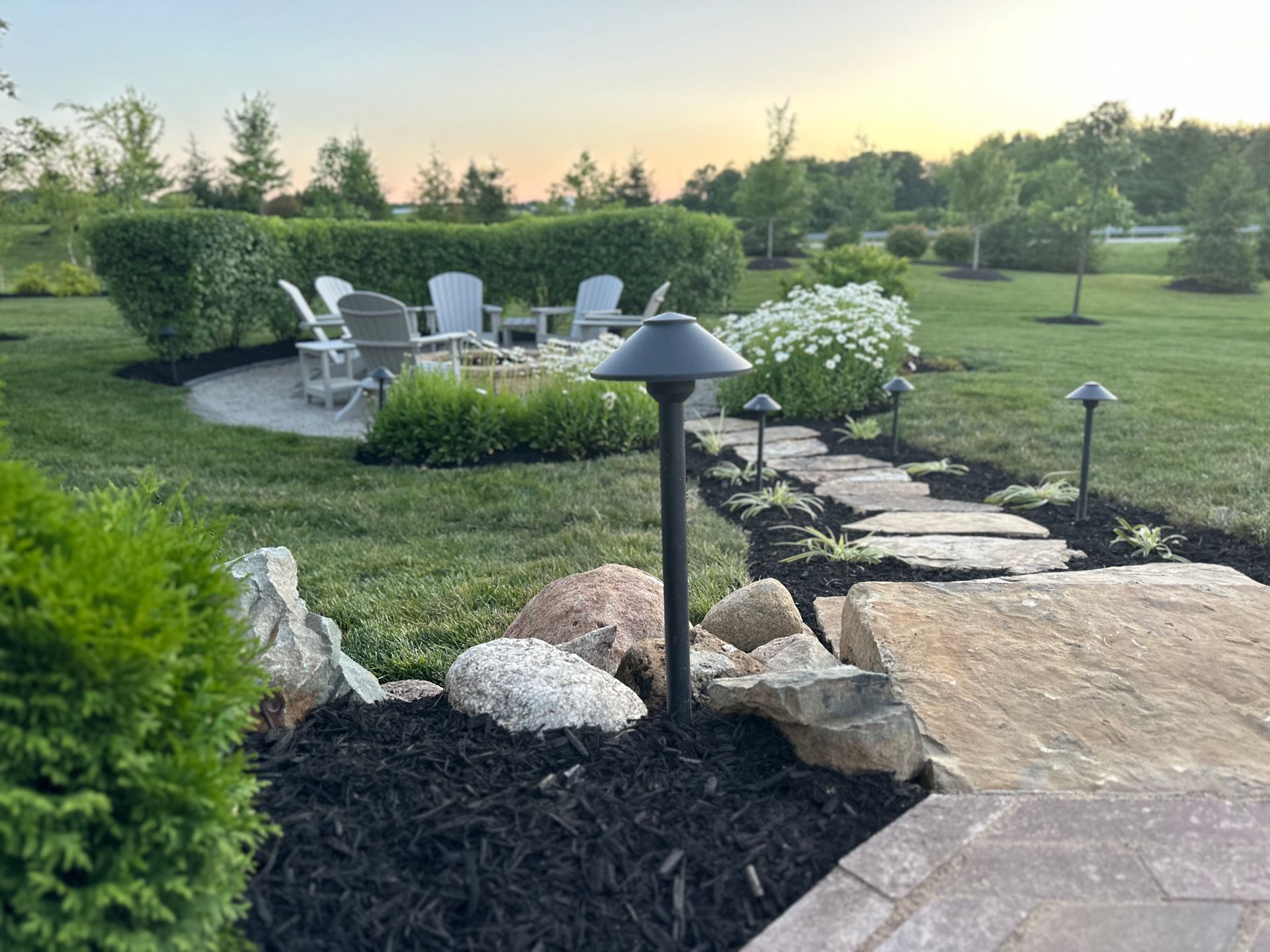 A sunset scene shows a stone path leading to a circular patio with Adirondack chairs, surrounded by hedges and landscaping.