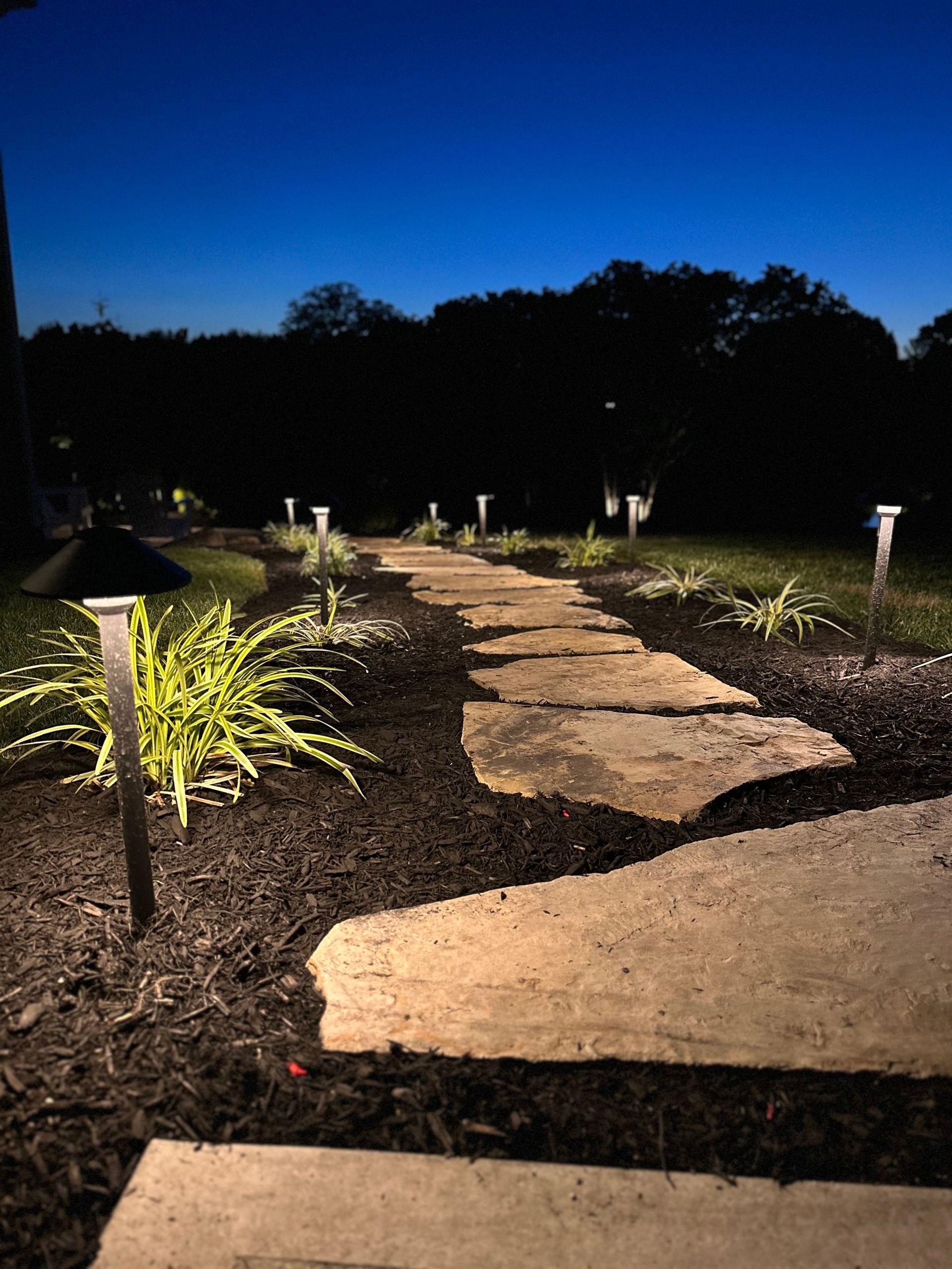 A stone pathway illuminated by small garden lights winds through a landscaped yard at twilight.