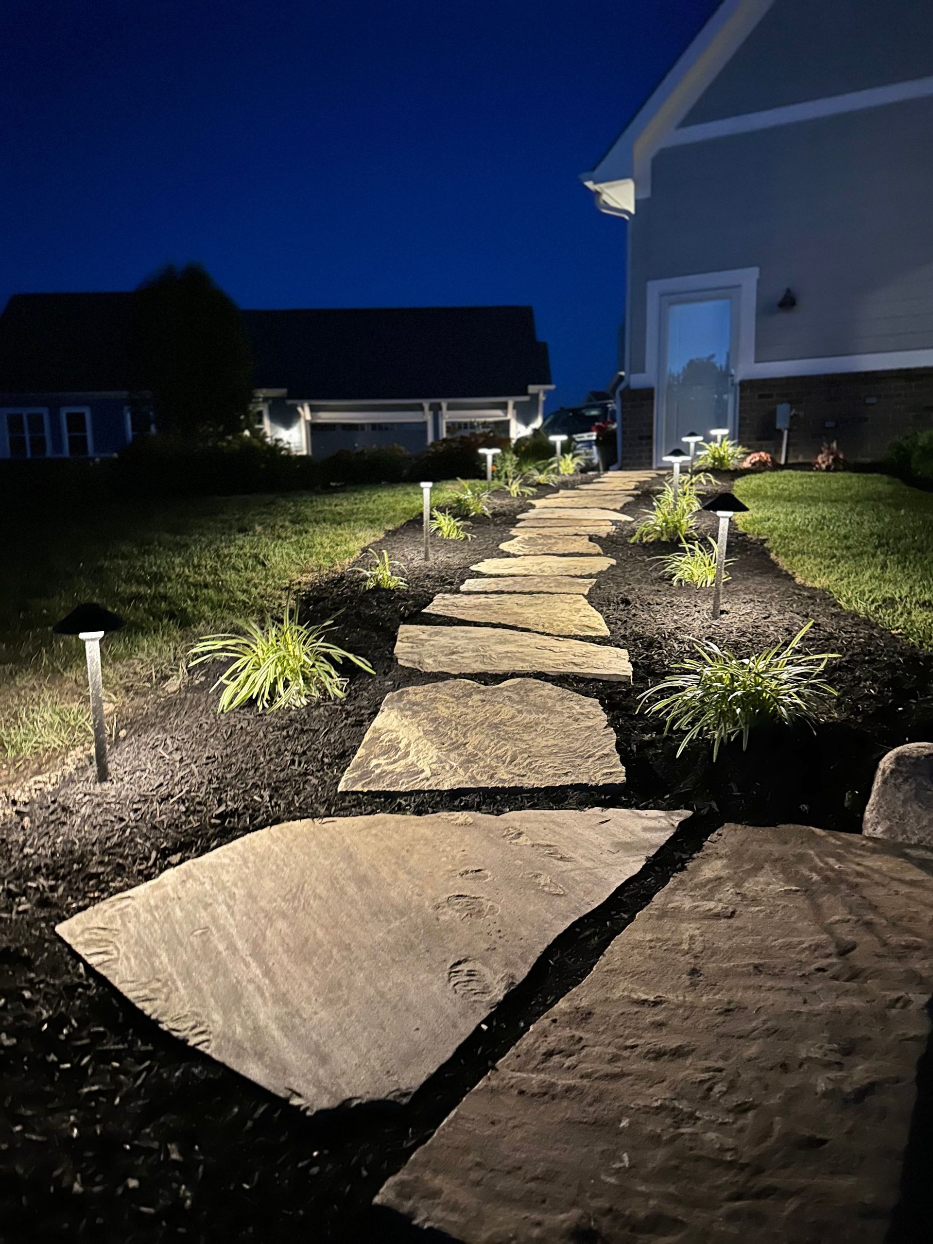 A stone garden path illuminated by solar path lights at dusk, leading toward a house.