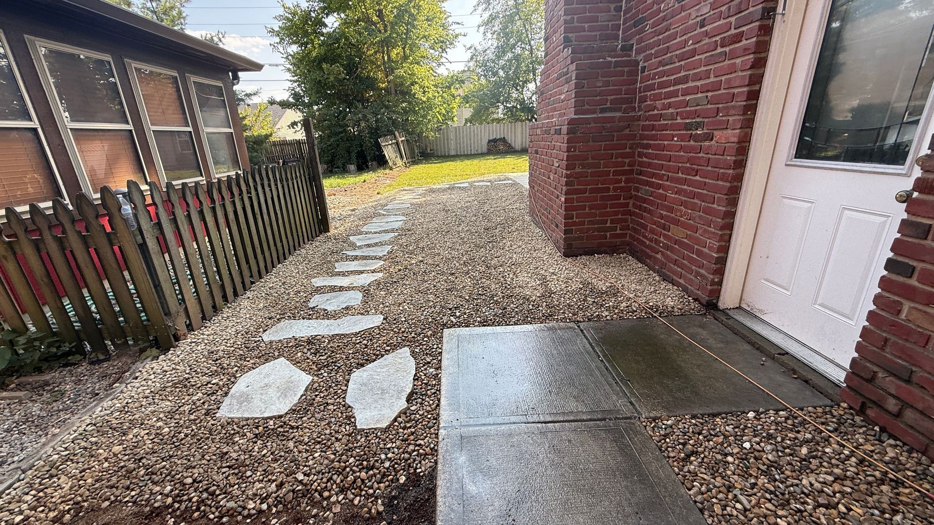 A stone path through gravel leads to a brick house entrance with a white door next to a brown picket fence.