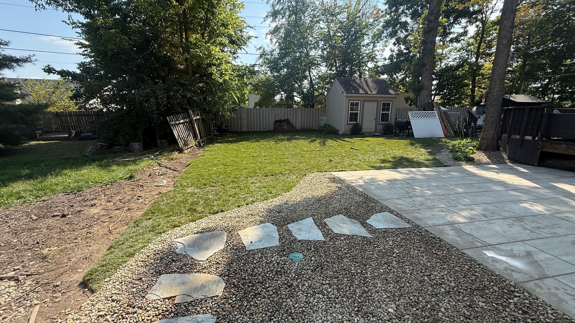 A backyard view featuring a gravel area with stone pavers, a patch of green grass, a small shed, and trees under a blue sky.
