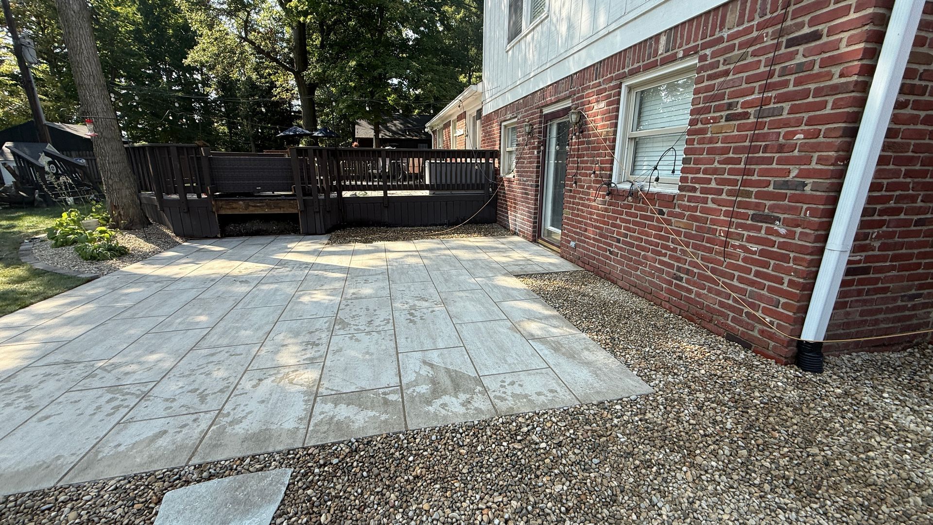 A stone patio and a gravel border next to a brick house with a raised wooden deck in the backyard.