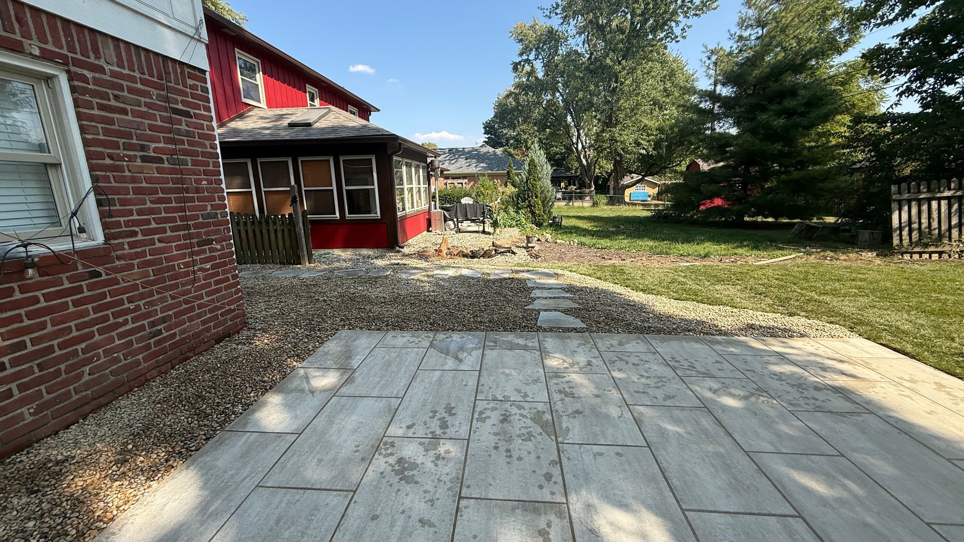 A backyard view featuring a red brick house wall, a stone patio, a red building addition, and a gravel-covered yard.