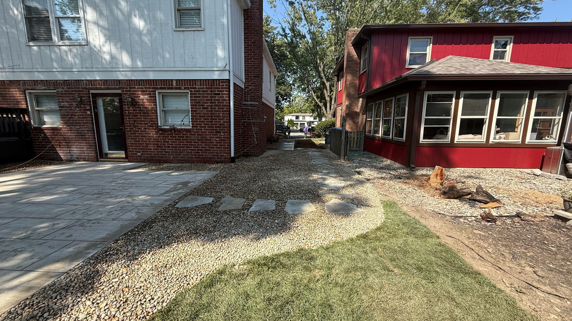 A white house and a red house sit side-by-side, separated by a gravel path and a patch of newly planted grass.