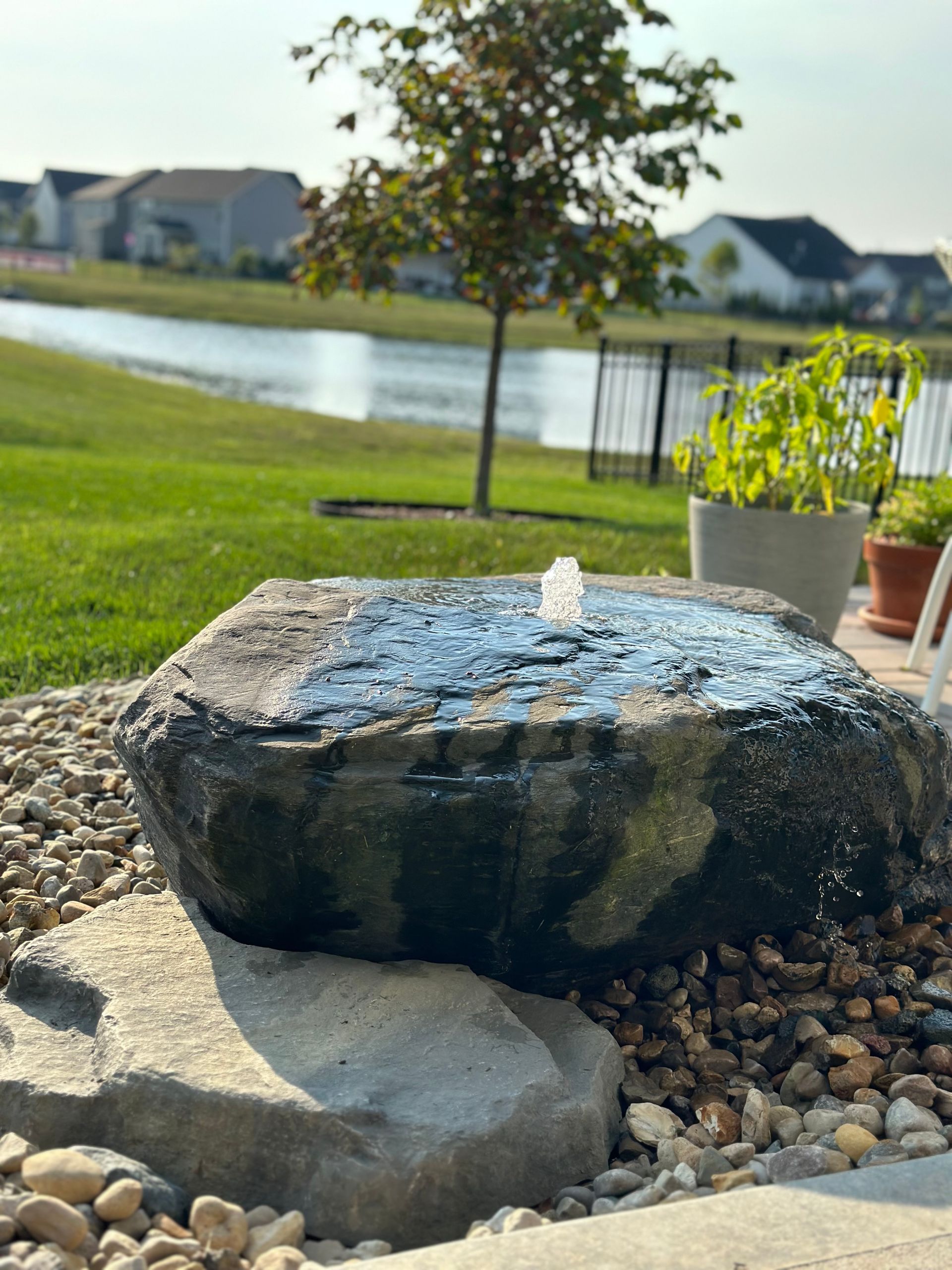 A backyard stone water feature fountain bubbler sits on a bed of gravel with a pond, tree, and houses in the background.