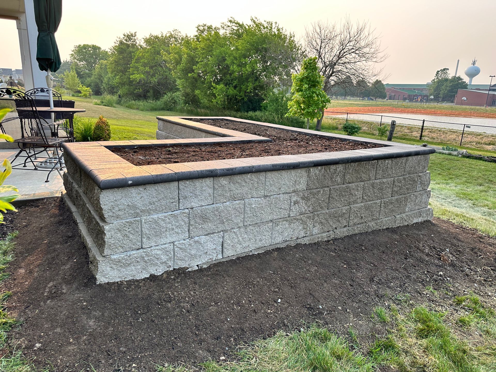 An L-shaped raised garden bed made of grey concrete blocks and filled with dark mulch in a grassy backyard setting.
