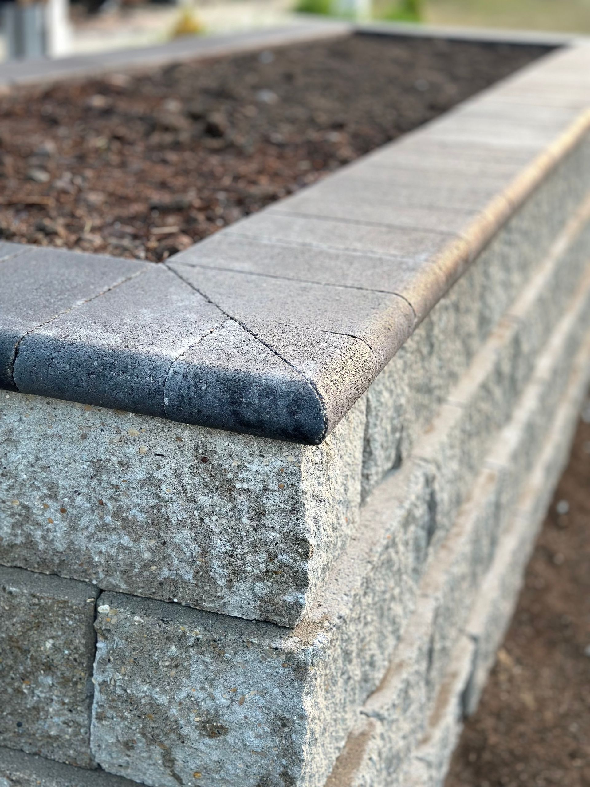 A raised garden bed constructed from grey, textured concrete blocks with smooth, angled capstones on the edges.