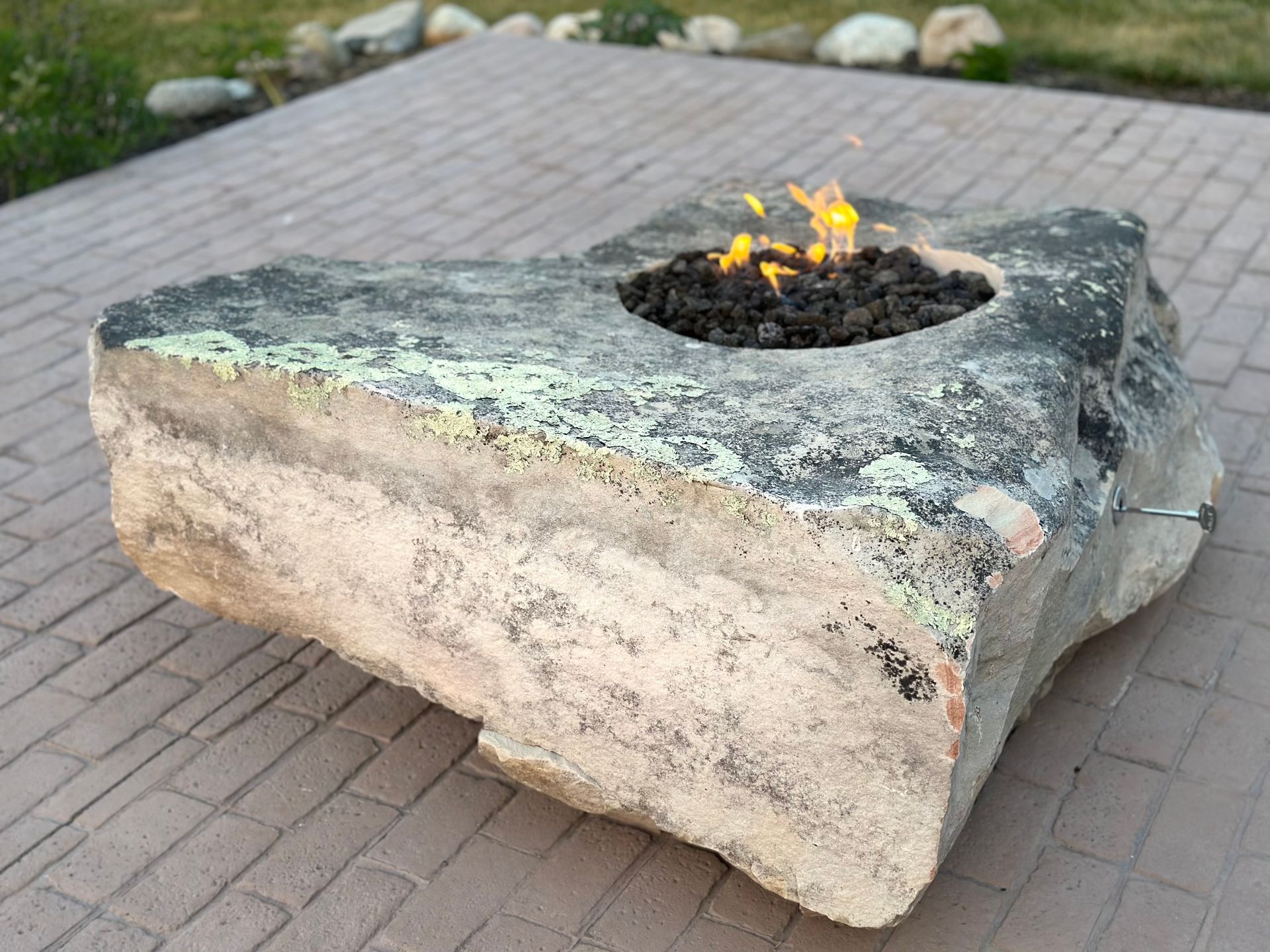 A large, natural-shaped stone fire pit with a small flame burning in a circular center depression on a paved patio.