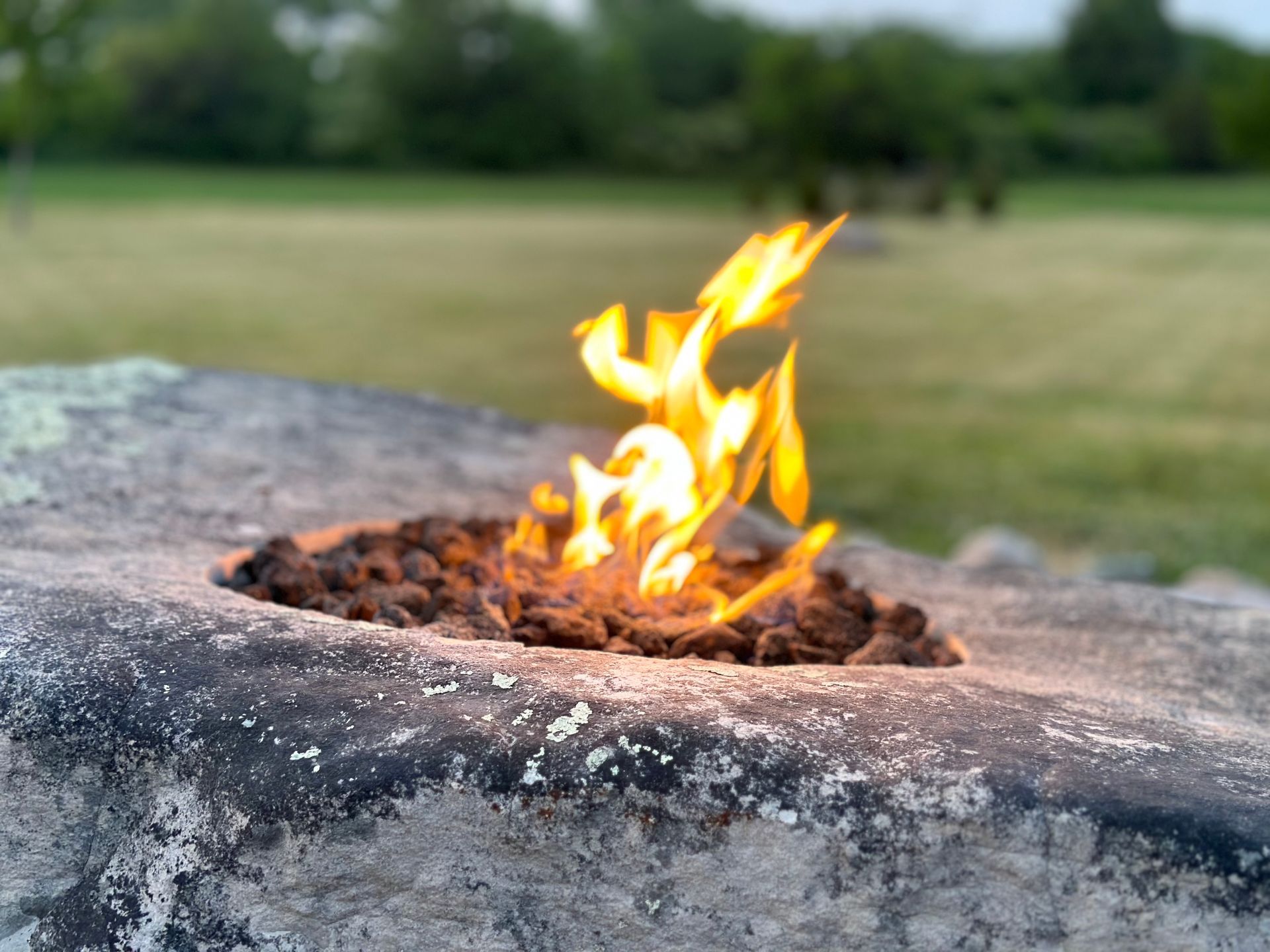 A small, bright yellow flame burns from a bed of dark volcanic rocks set into a textured, weathered stone outdoors.