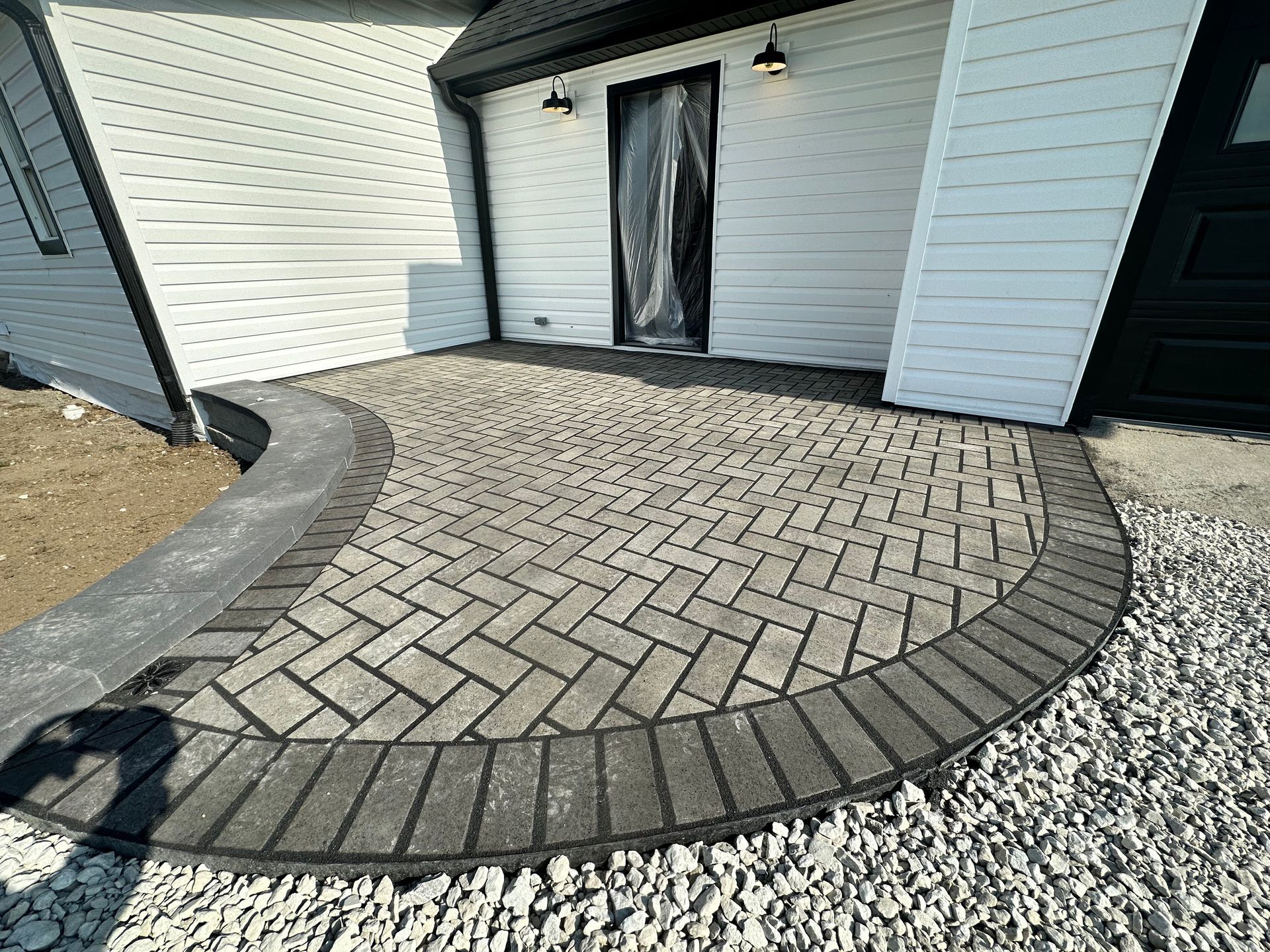 A newly installed gray herringbone brick patio with a dark charcoal curved border edge in front of a white house.