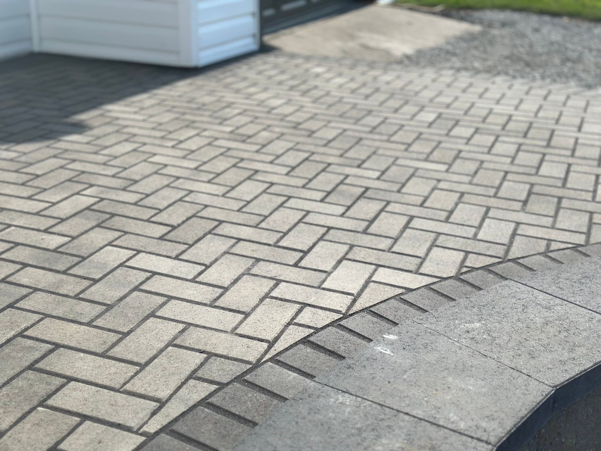 A gray herringbone-patterned stone patio with a curved border of darker rectangular pavers leading to a white wall.
