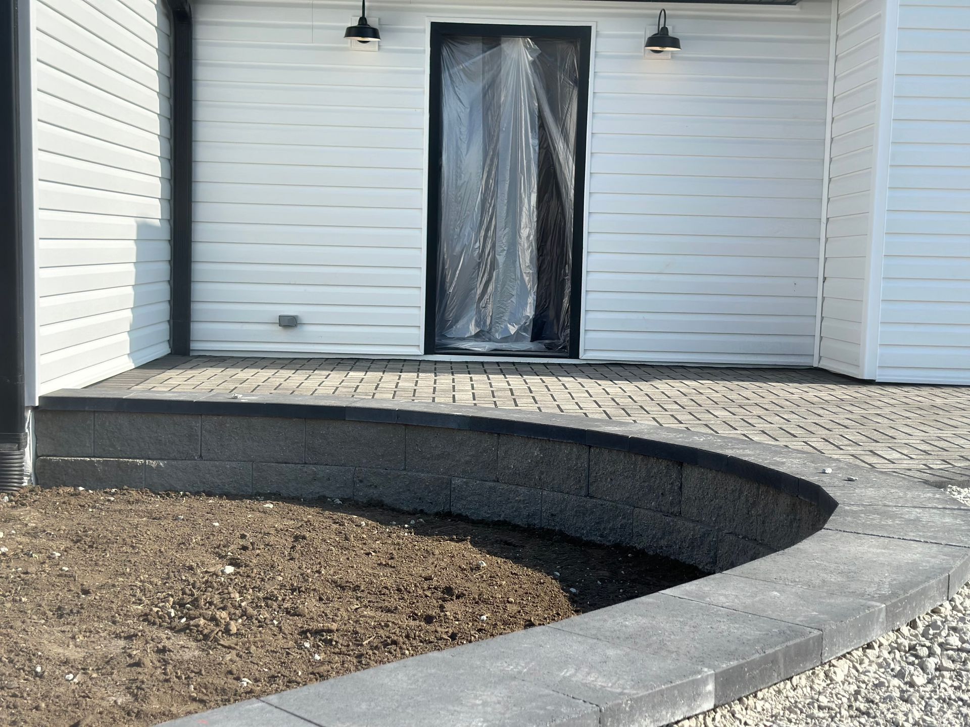White siding house with a paver patio and a curved gray stone retaining wall bordering an area of soil.