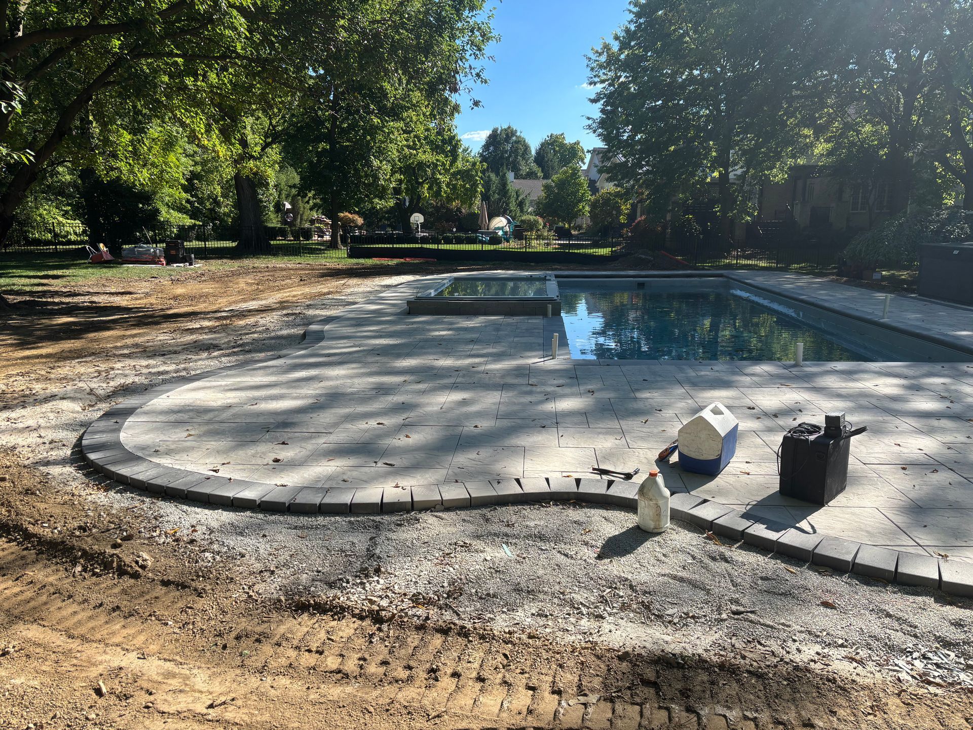 A sunlit backyard features a rectangular swimming pool with a new stone paver patio and a partially landscaped dirt area.