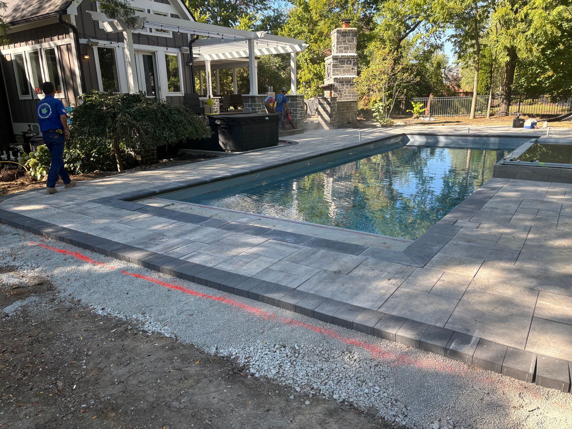 A person walking beside a newly installed swimming pool with a stone patio, surrounded by trees and a house.