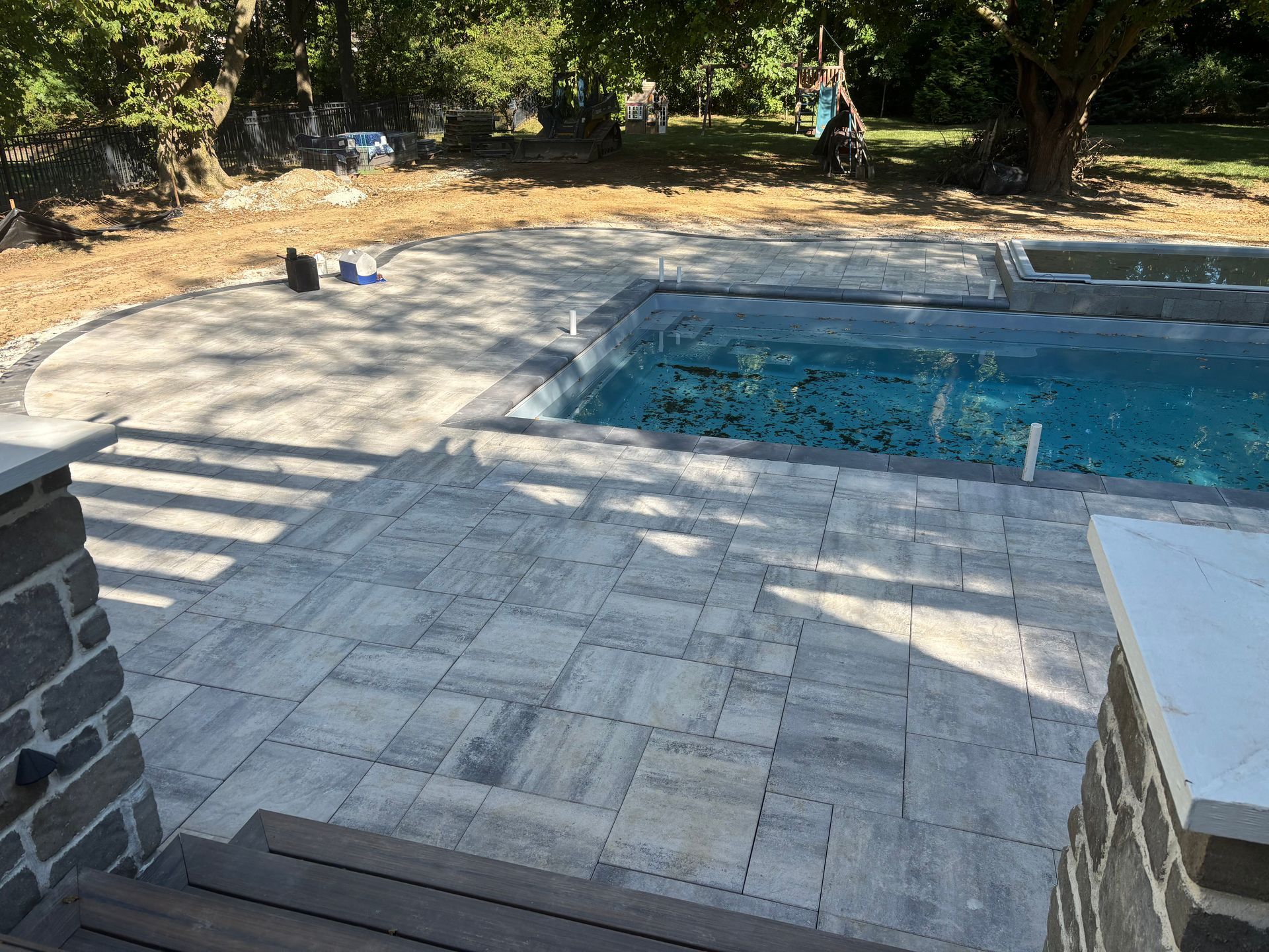 A light-gray paver patio leads to a blue swimming pool in a sunny, tree-lined backyard setting.