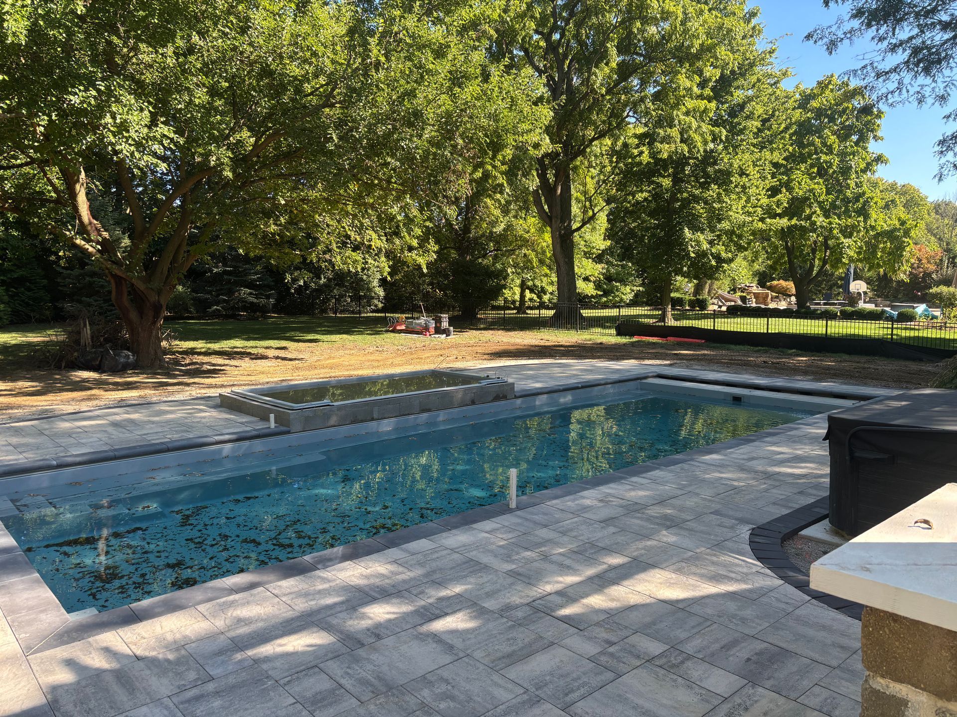 An outdoor rectangular swimming pool surrounded by stone pavers, set against a backdrop of lush green trees on a sunny day.