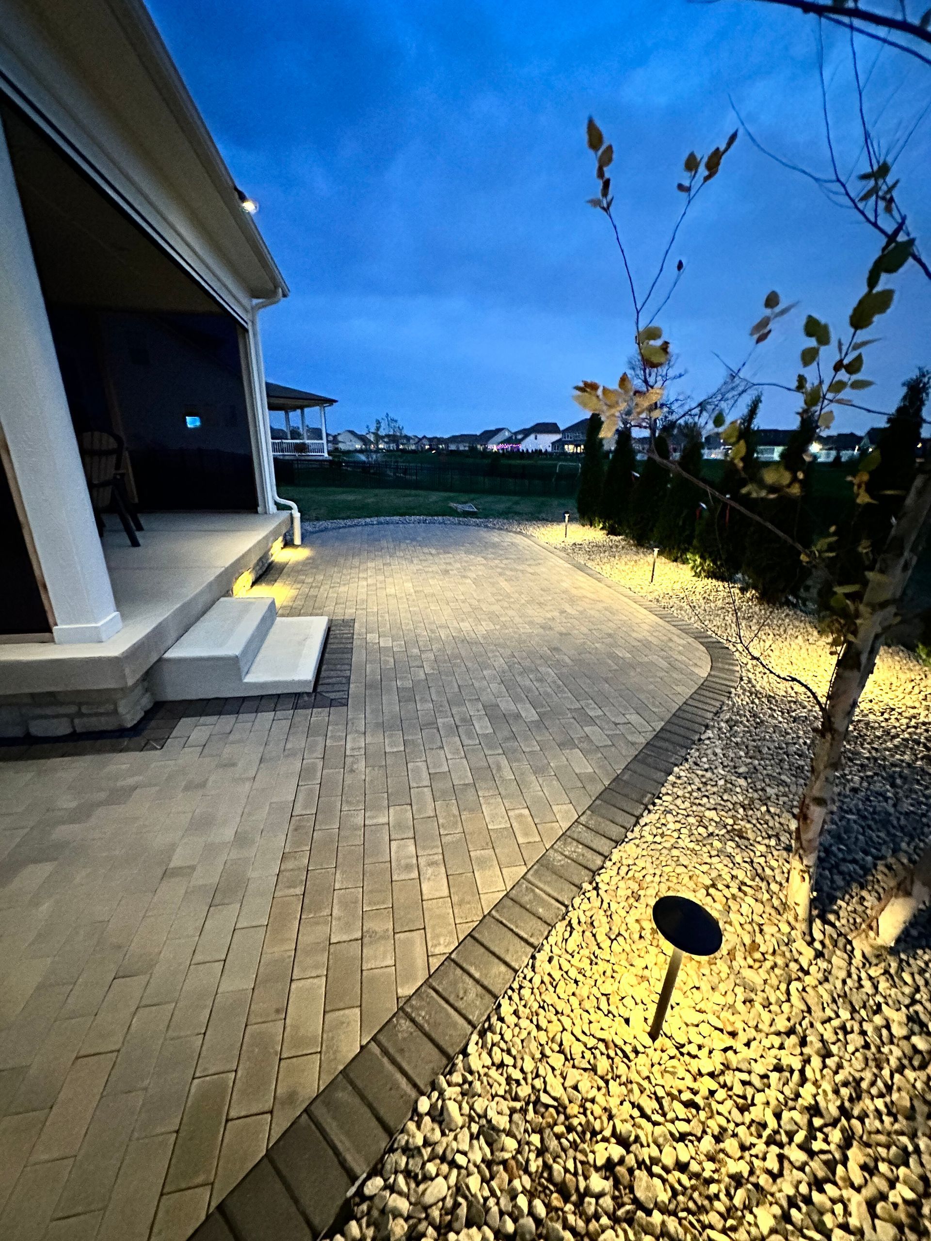 A patio made of light-colored bricks at dusk, illuminated by glowing path lights, bordering a rock garden with a small tree.