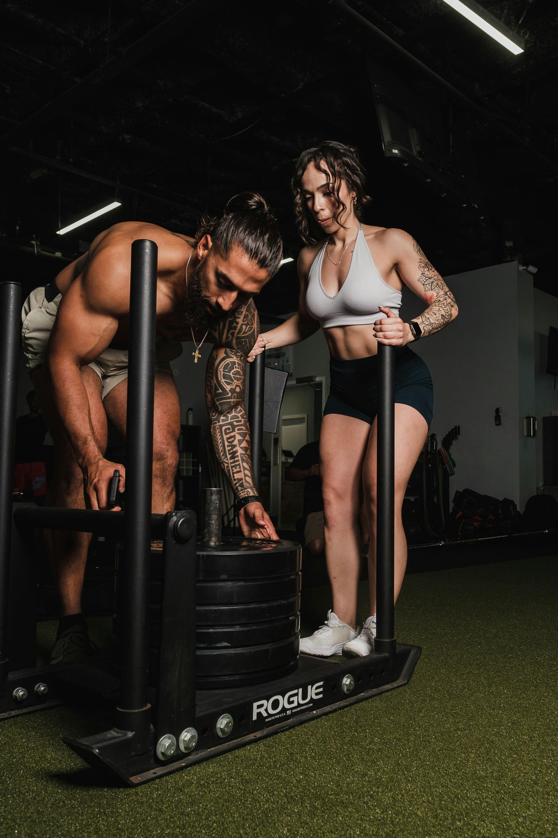Vrouw gebruikt een fitnessapparaat onder begeleiding van een trainer in een sportschool, met zwarte en oranje accenten.