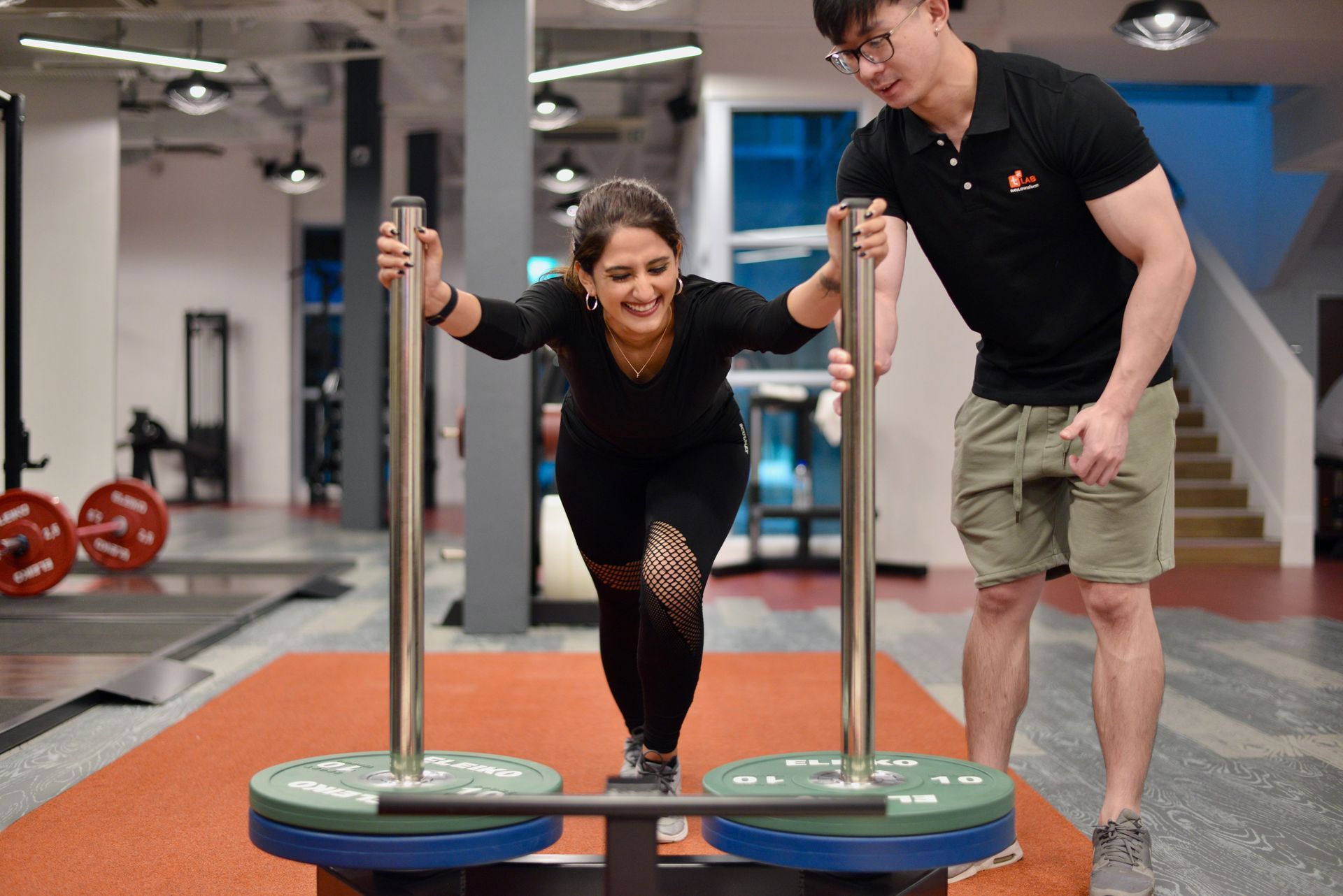 Twee mensen duwen een verzwaarde slee in een sportschool; een van hen lacht terwijl ze aan het sporten zijn.