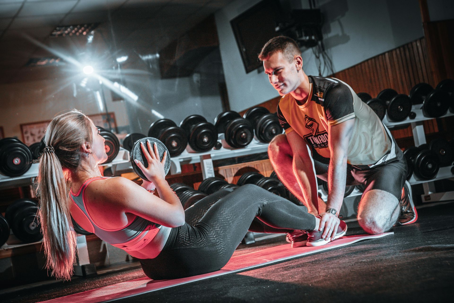 Een vrouw doet lat pulldowns met een trainer, zittend in een sportschool. Ze dragen rode kleding. 