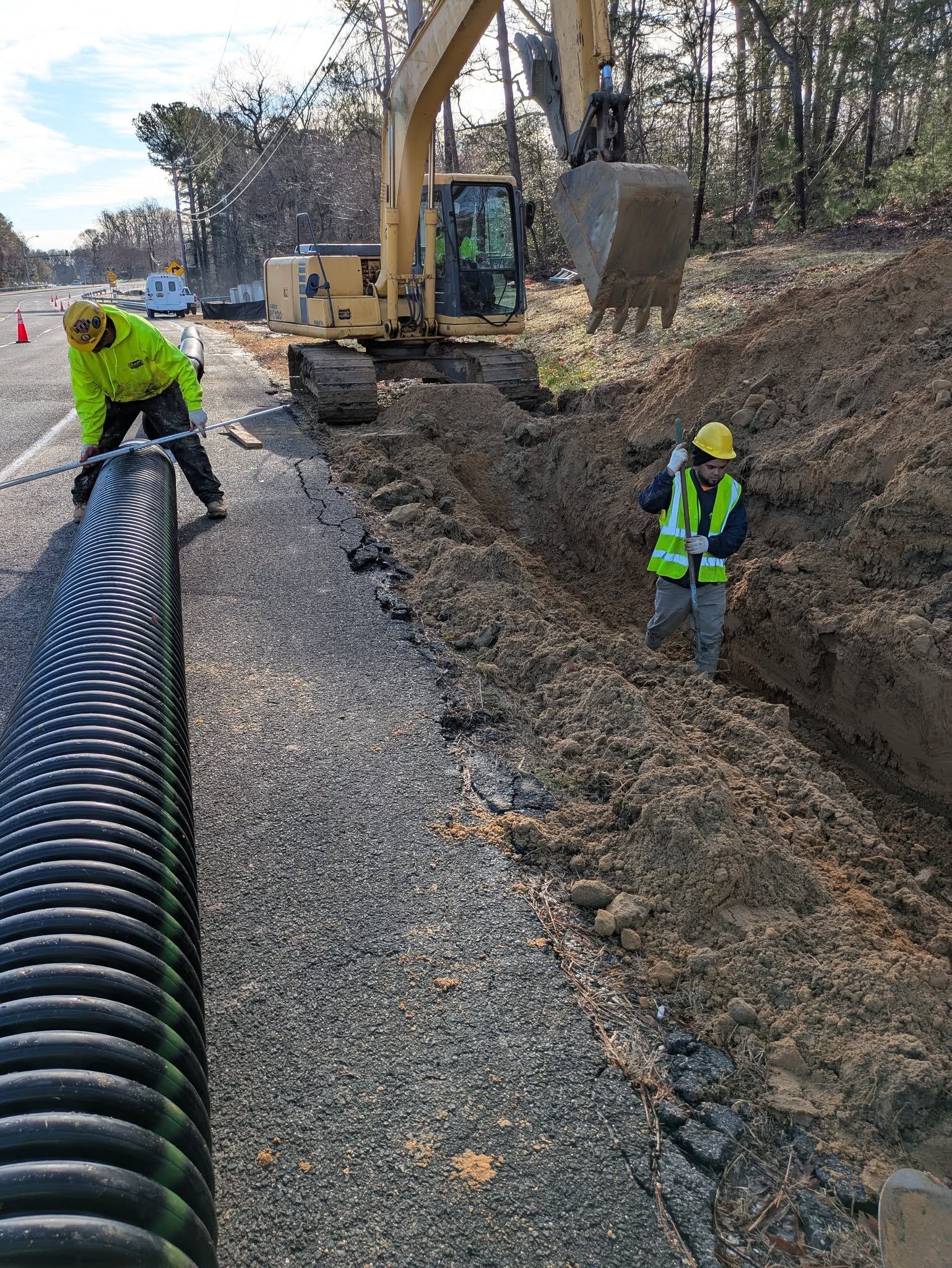 Two workers in high-visibility gear install a long, black corrugated pipe next to a trench being dug by an excavator.
