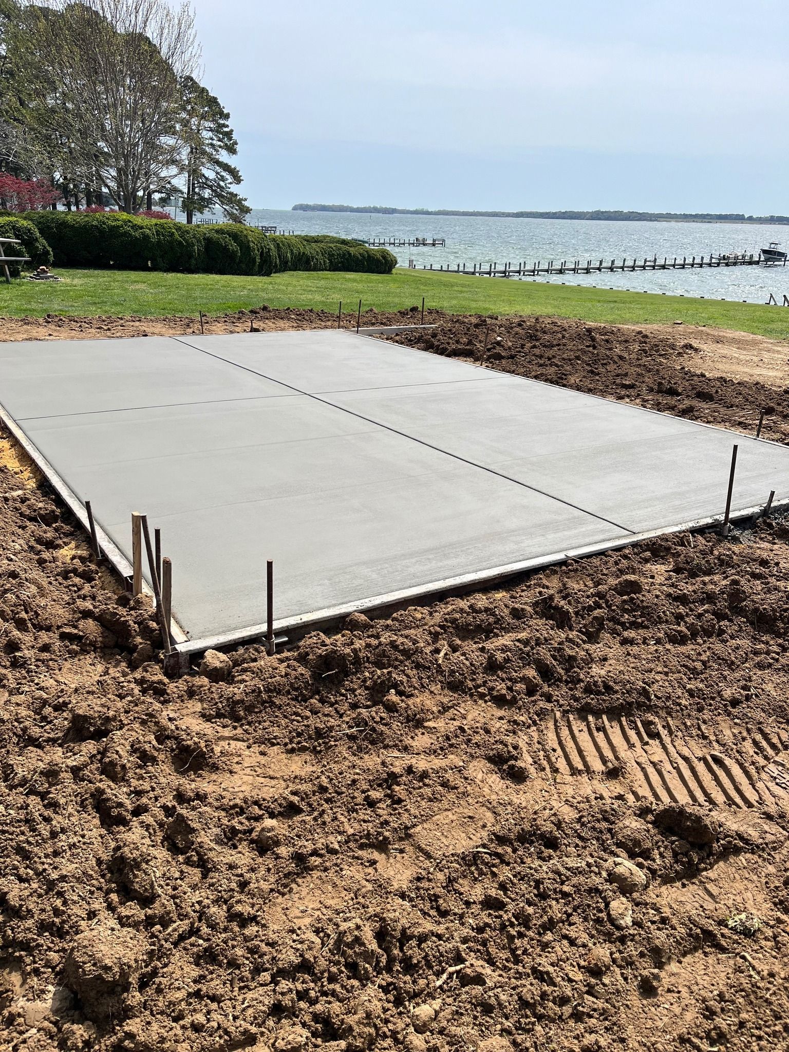 A fresh rectangular concrete slab sits on cleared dirt near a grassy waterfront under a clear sky.