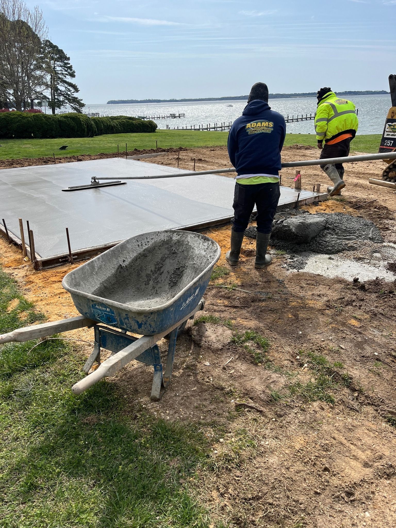 Two workers smooth fresh concrete for a patio near a waterfront, with a wheelbarrow in the foreground.