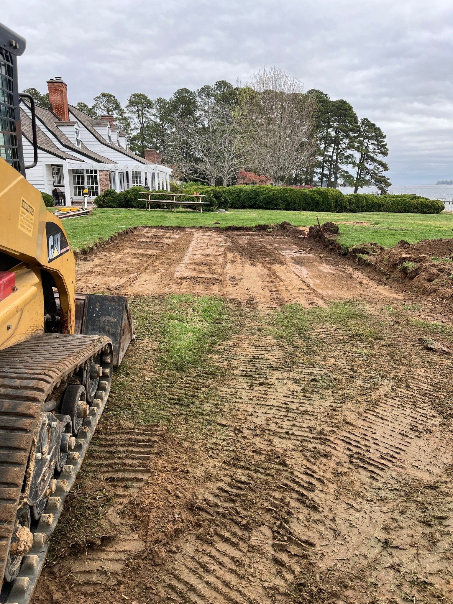 A yellow skid steer loader sits on a muddy, excavated dirt path leading toward a white house with a porch.