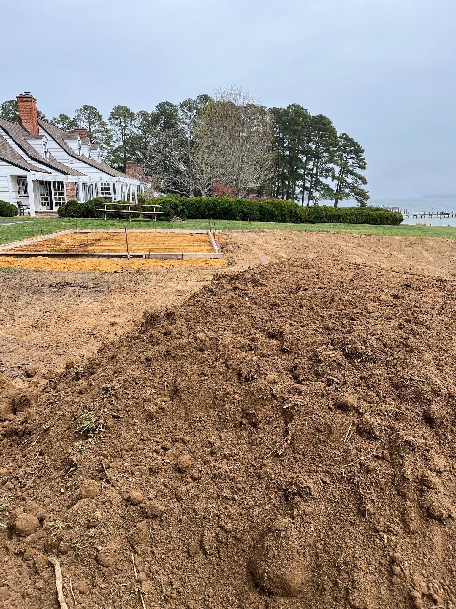 A landscape view of freshly tilled soil in a large garden plot near a house, trees, and a body of water.