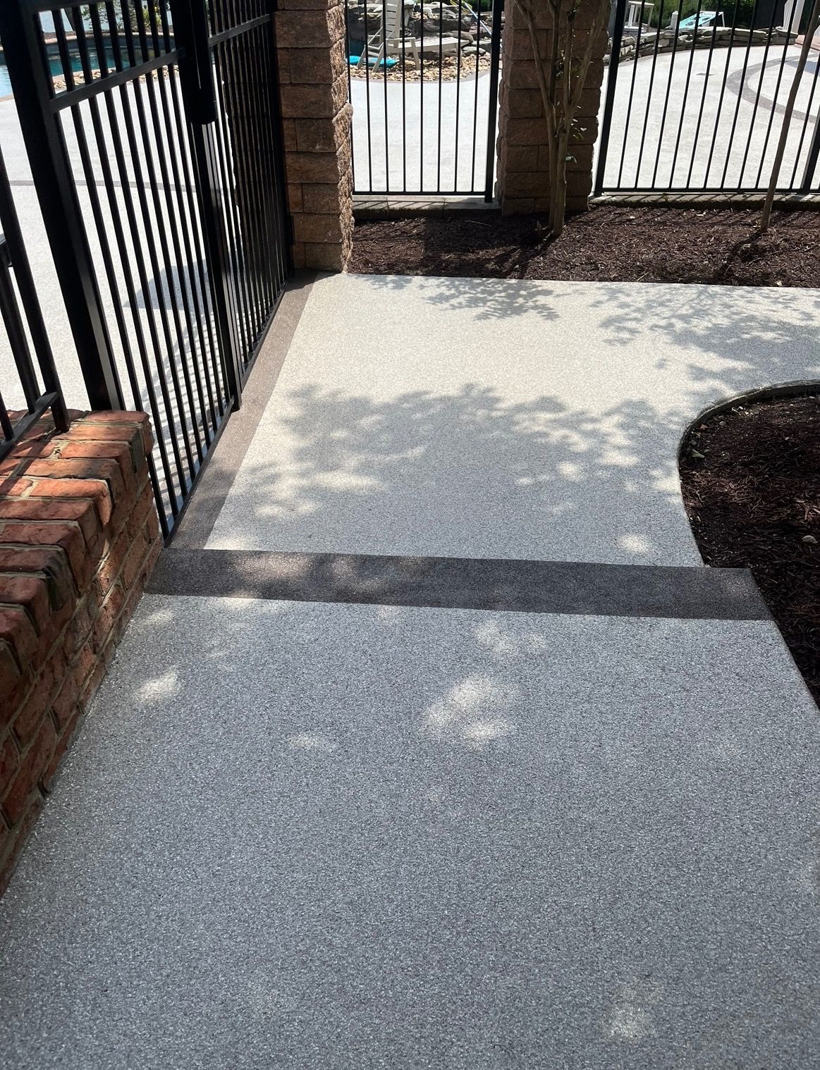 Two concrete steps with a textured, gray aggregate finish lead to a black metal gate and brick pillar outdoors.