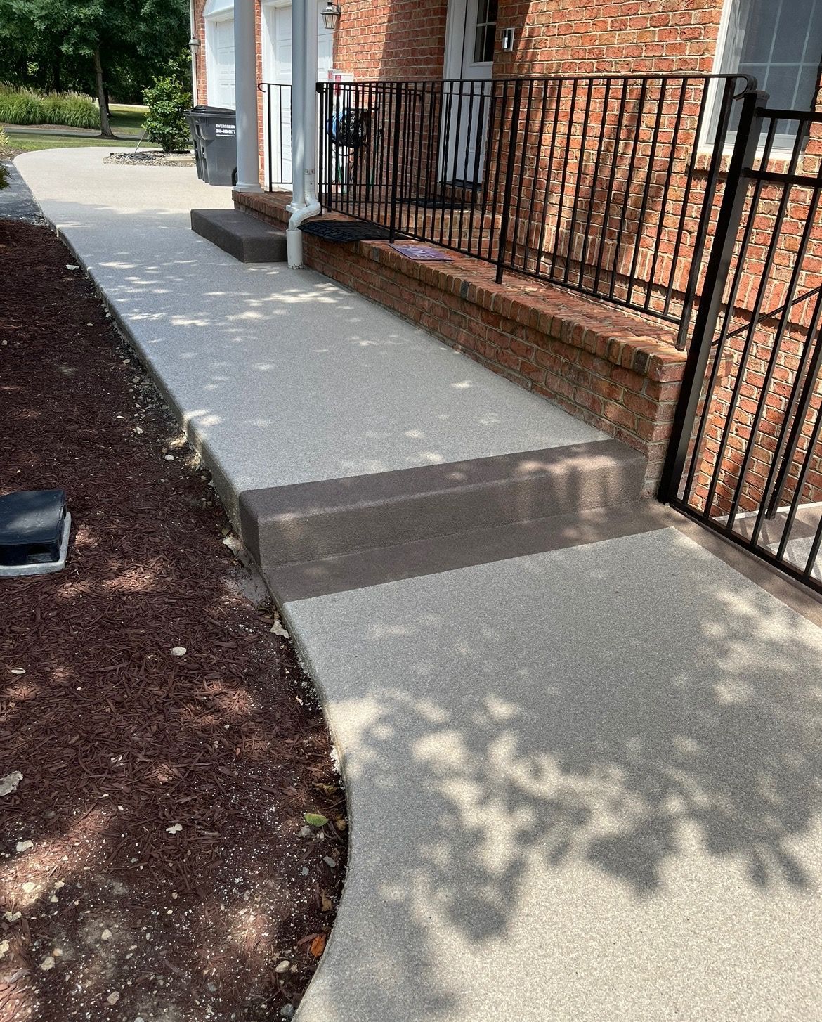 A concrete pathway leading to a brick building, featuring a small raised step and a black metal railing.