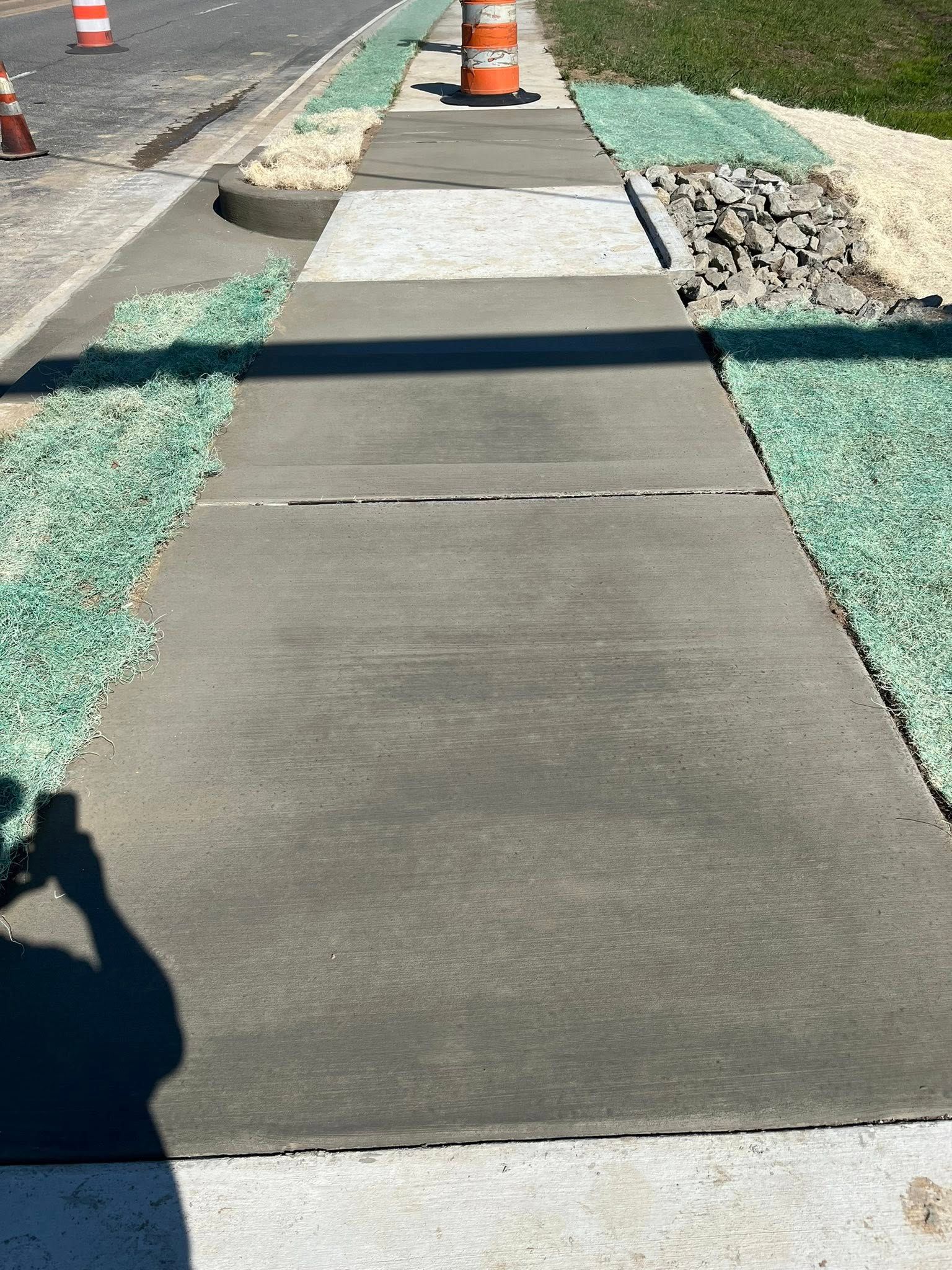 A concrete sidewalk under construction, flanked by green ground cover and gravel, with orange traffic cones in the distance.
