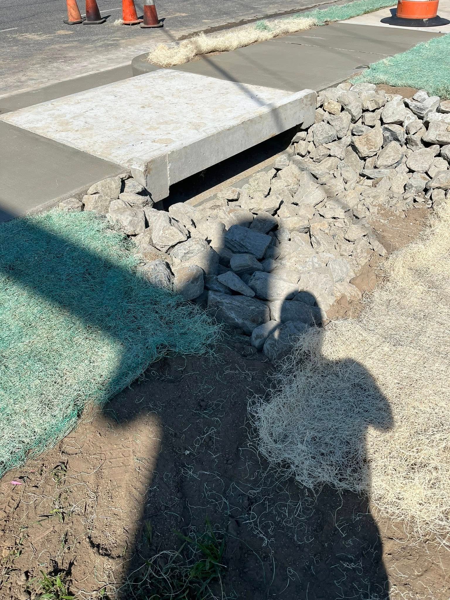 A concrete storm drain entrance surrounded by patches of green, tan, and grey landscaping gravel on a roadside.