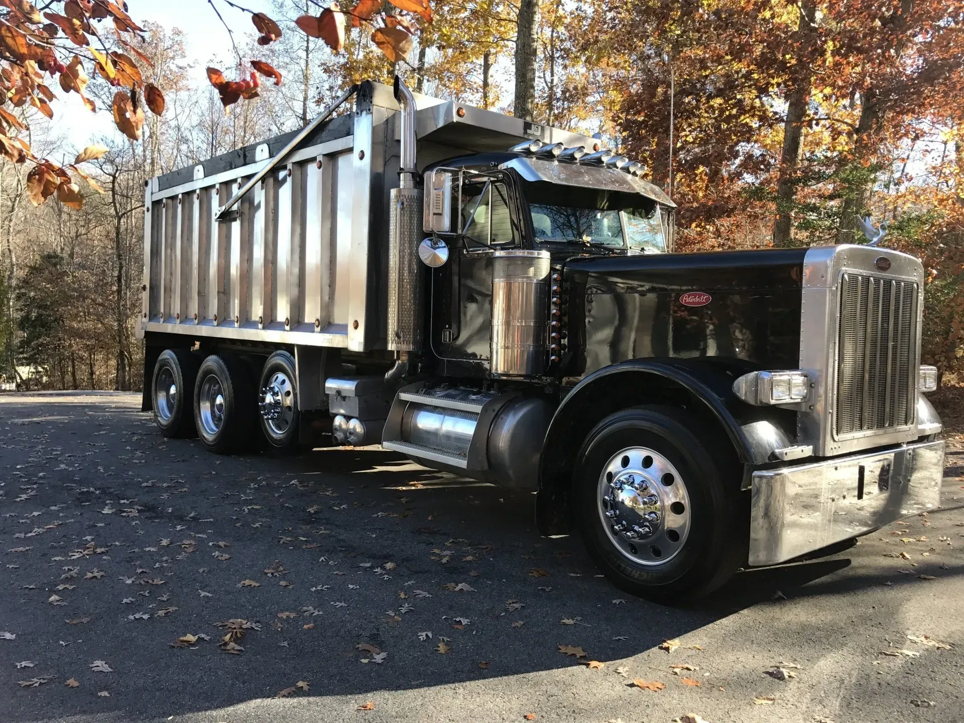 A shiny, black dump truck parked on an asphalt road surrounded by trees with autumn leaves.